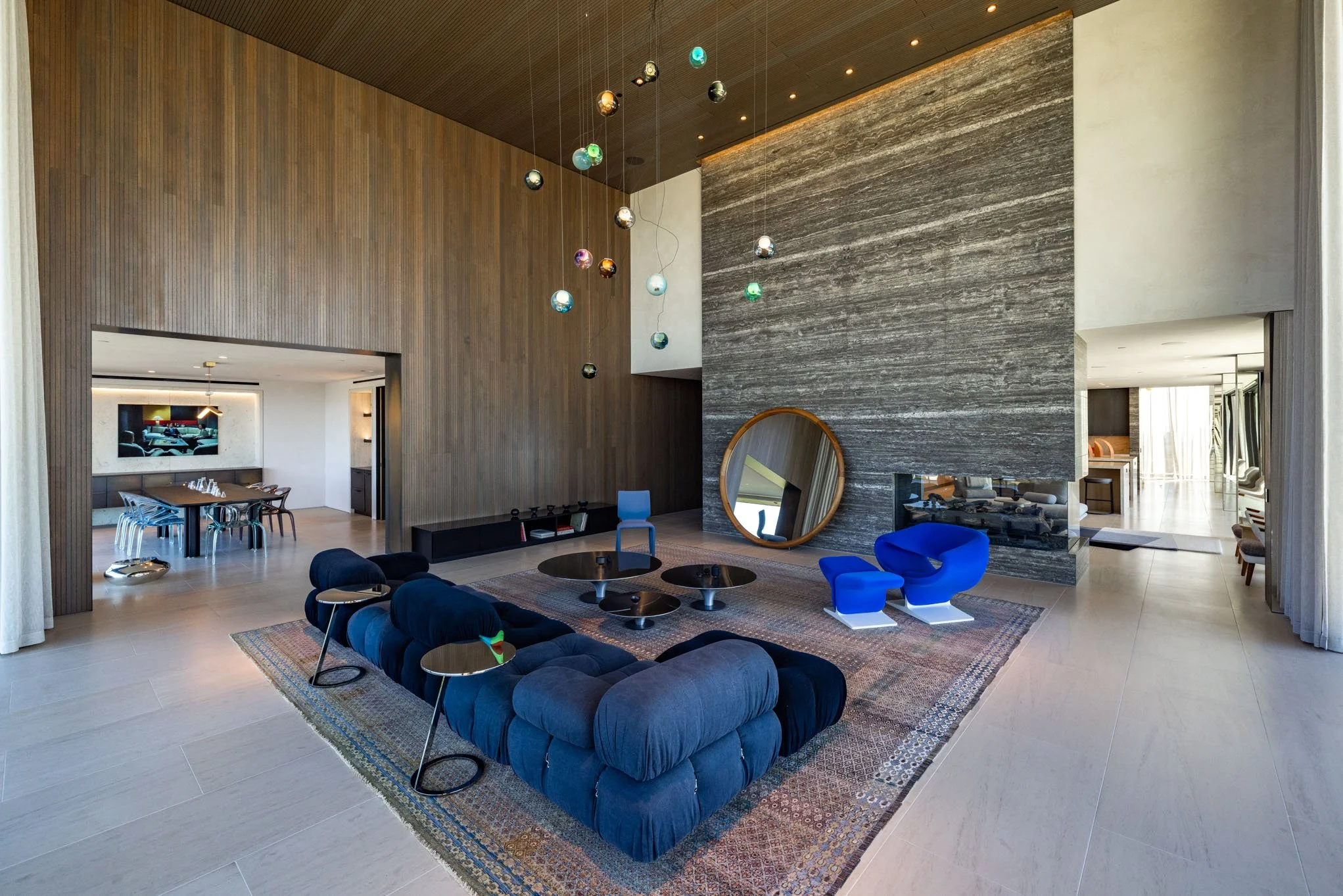 Spacious modern living room with textured wood and stone walls, blue velvet seating, and artistic pendant lighting