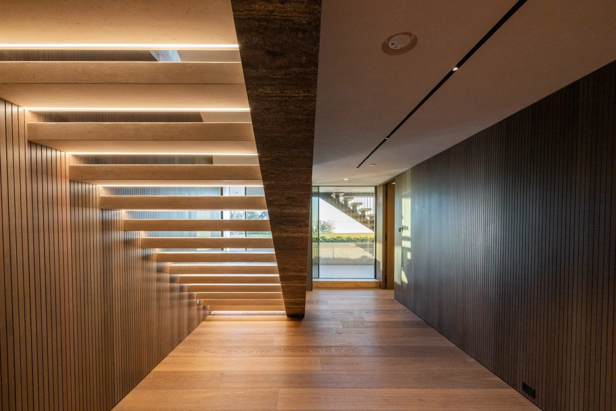 Modern interior hallway with wooden flooring, vertical wood-paneled walls, and a staircase with horizontal wooden steps and lighting. At the end, there's a glass door and stairs outside.