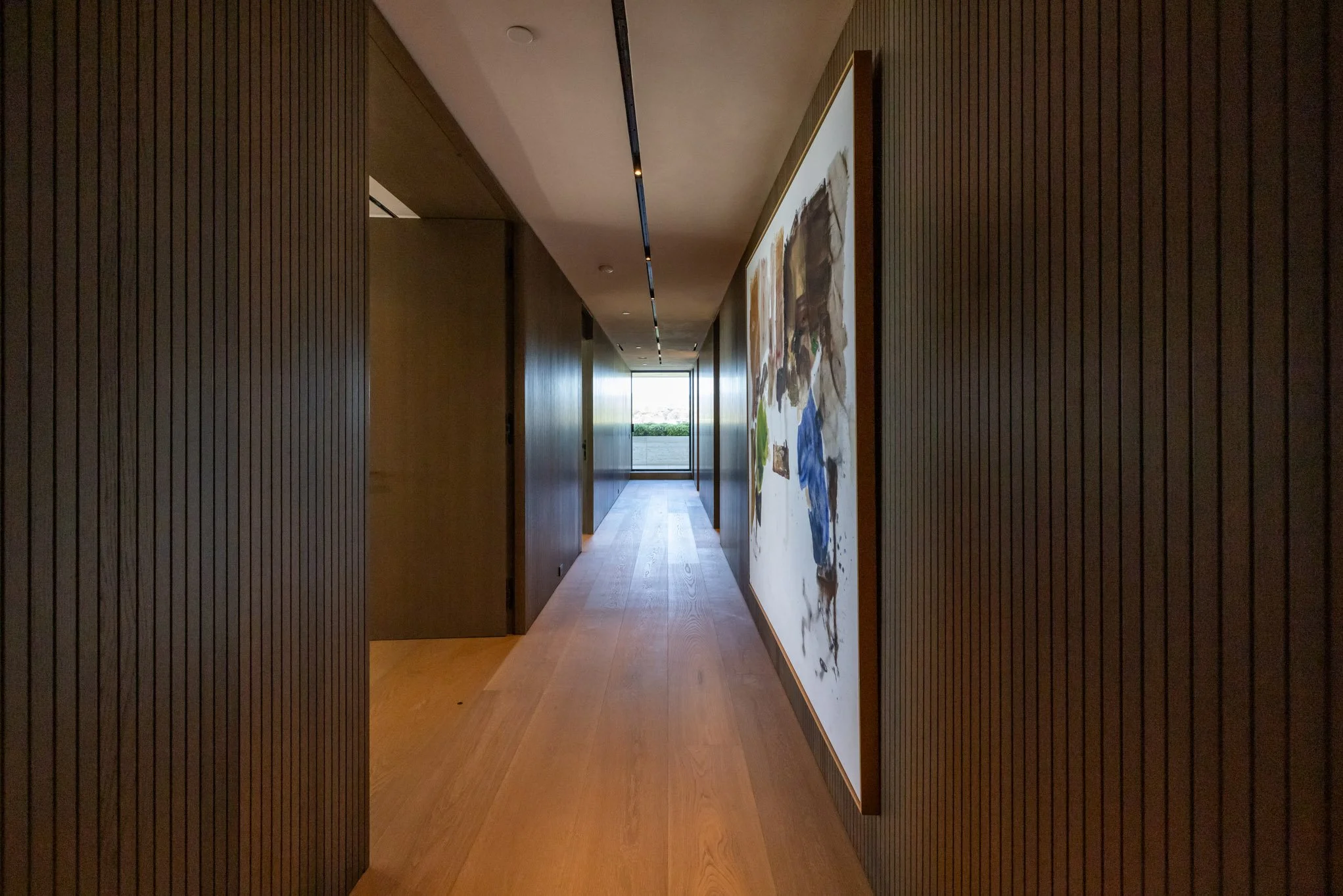 Long corridor with dark wooden paneled walls, a wooden floor, and an abstract painting on the right wall. Natural light from a window at the end of the hallway.