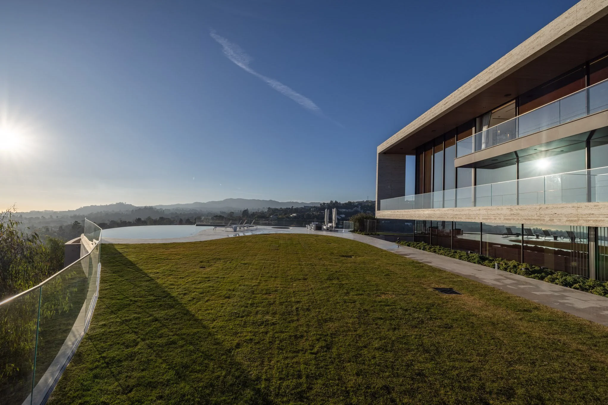 Modern house with large glass windows and balcony overlooking a grassy lawn, overlooking a scenic view of hills and mountains, with the sun shining in a clear blue sky.