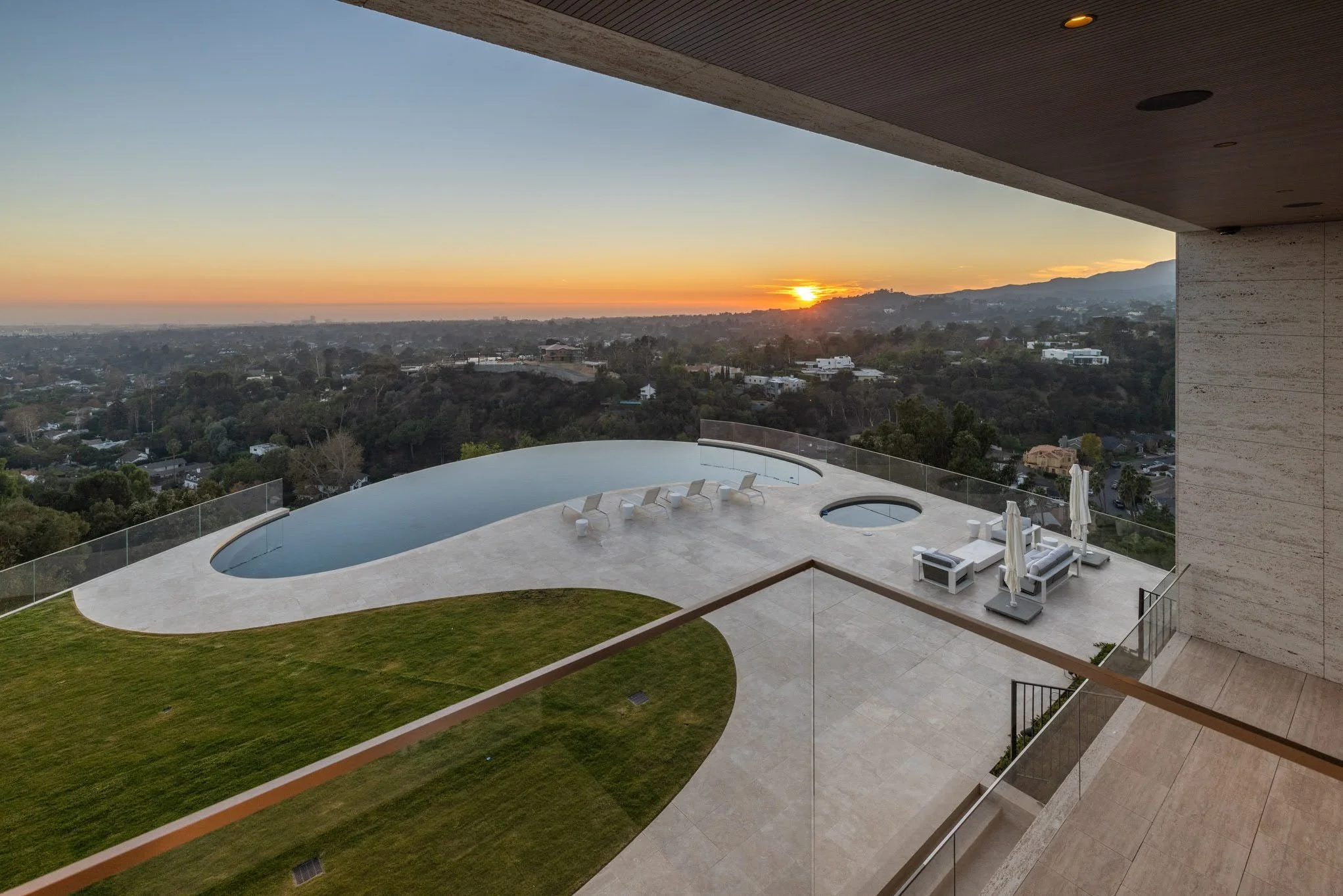 Luxury balcony with a curved swimming pool, outdoor lounge chairs, umbrellas, and a hot tub overlooking a scenic landscape at sunset.