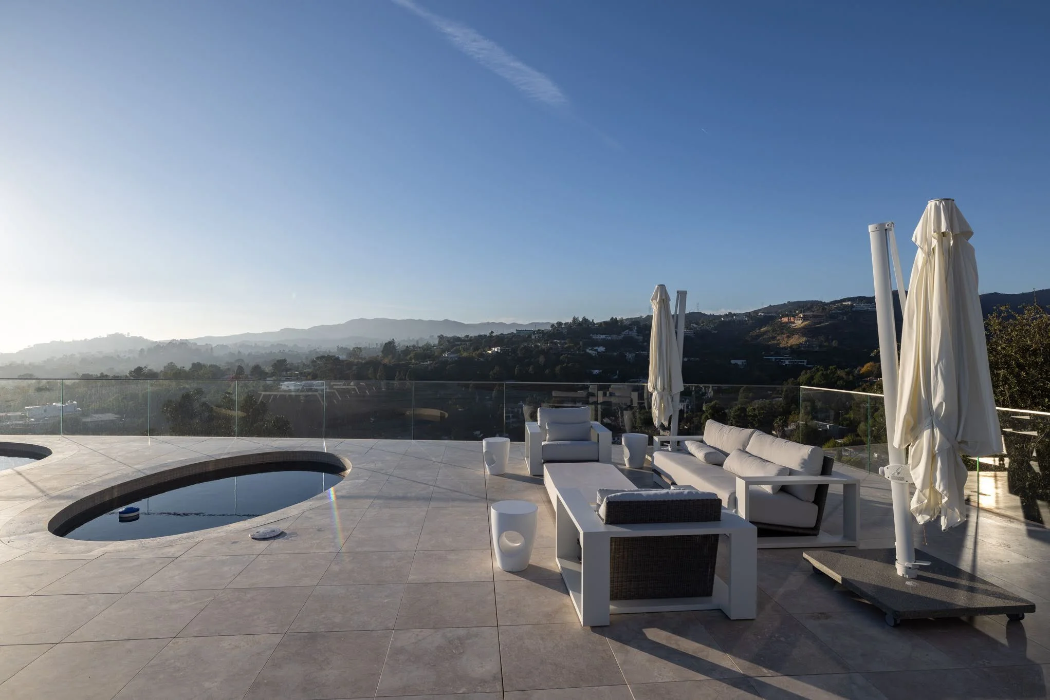 Outdoor rooftop patio with white lounge furniture, umbrellas, and a jacuzzi overlooking a scenic hilly landscape at sunrise or sunset.