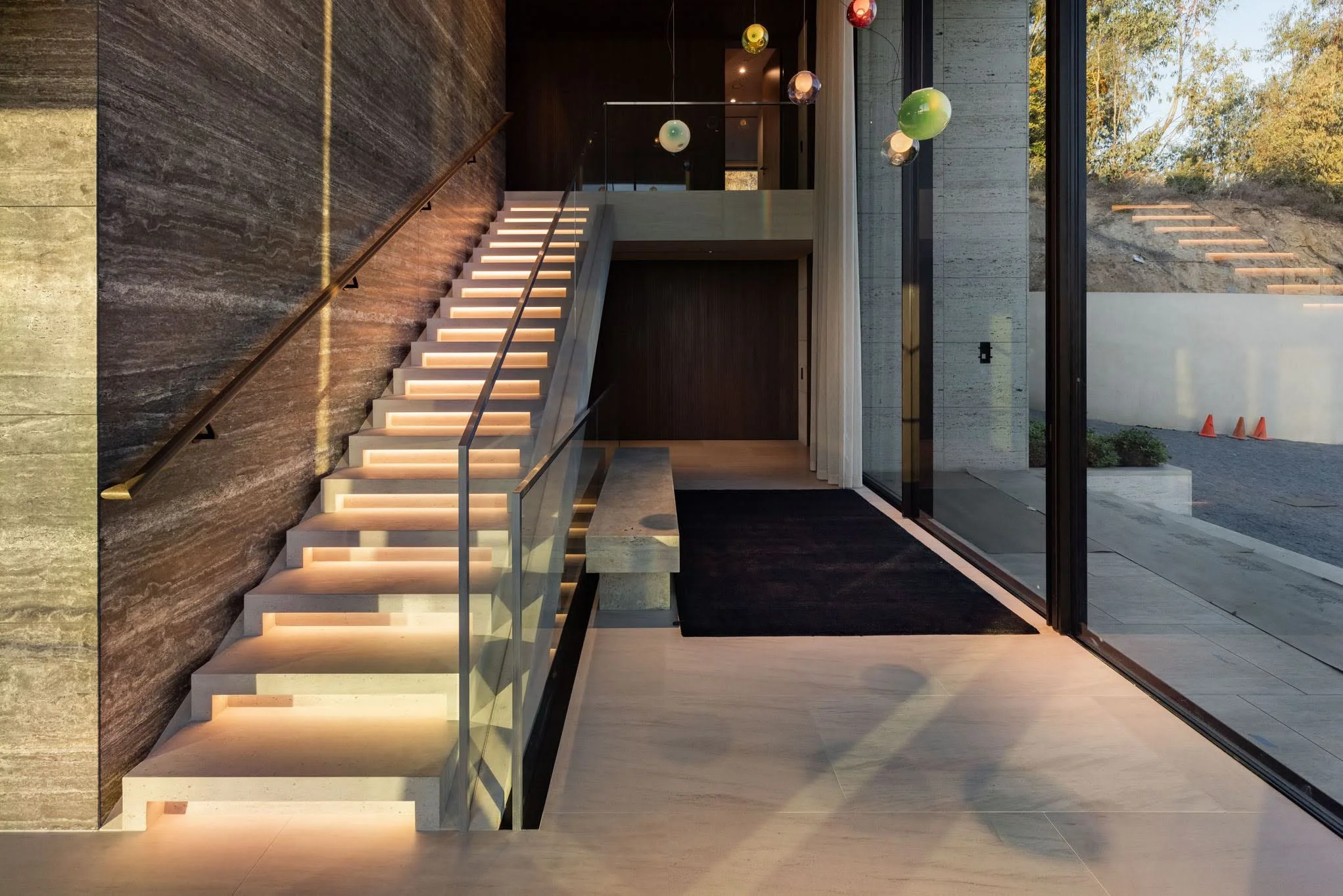 Modern interior with illuminated staircase, dark and light textured walls, large glass doors, and hanging spherical pendant lights.