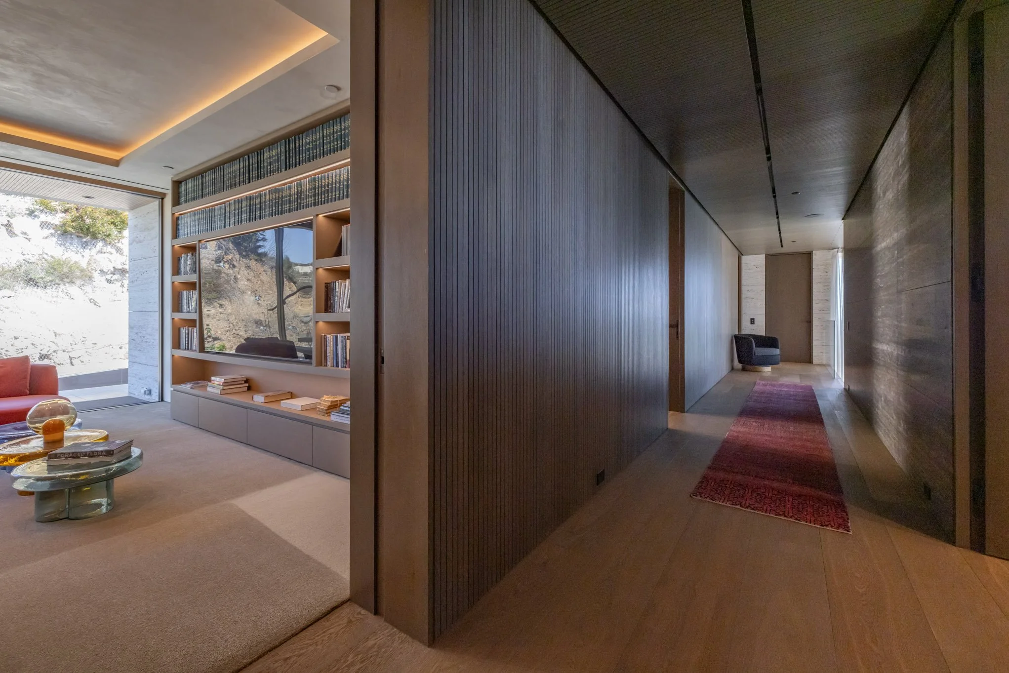 Interior view of a modern house with a spacious living room and hallway, featuring wooden floors, a large window with a view of nature, and minimalist furniture including a bookshelf, a sofa, and a coffee table.