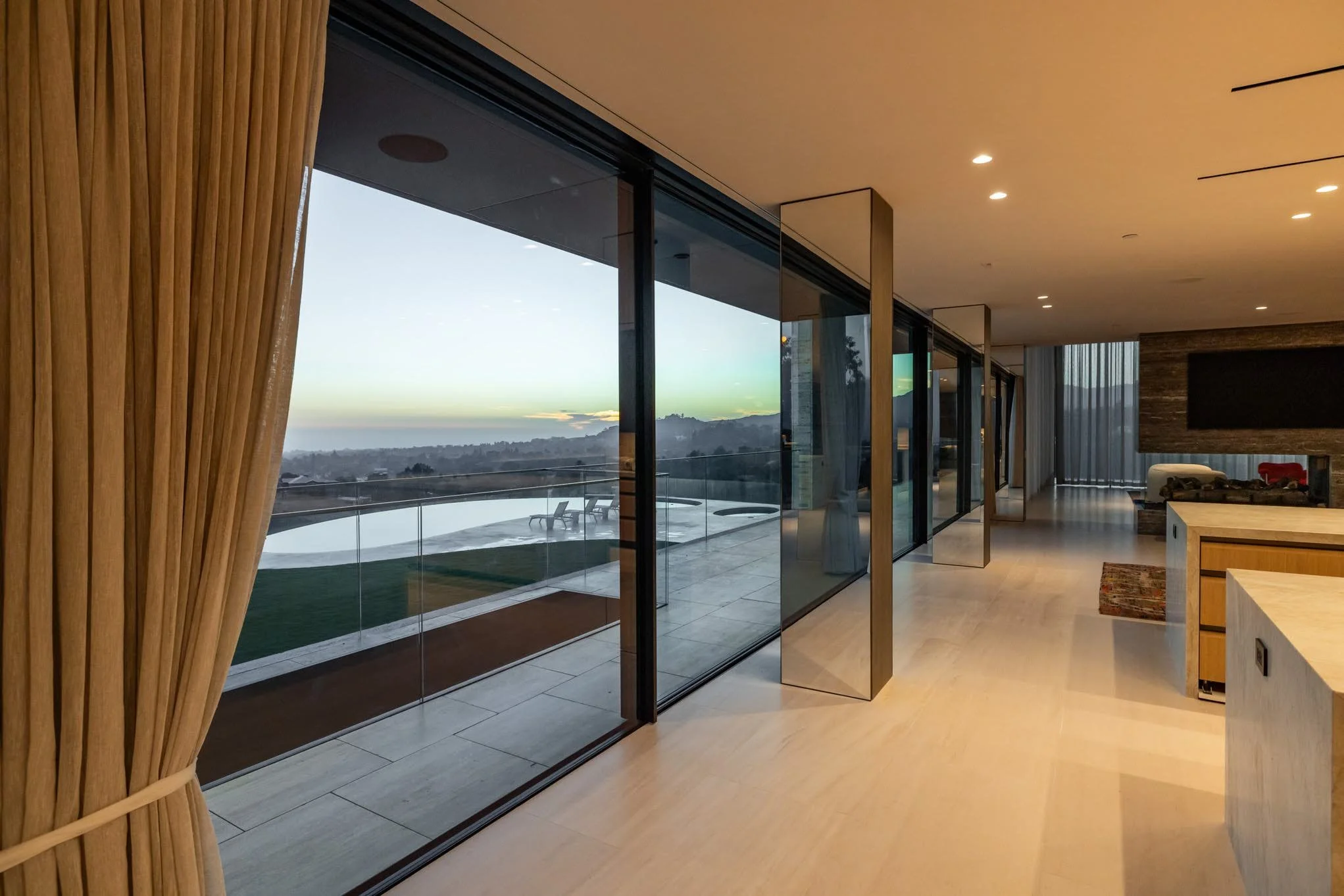 Modern living room with floor-to-ceiling glass windows showing an outdoor patio and scenic landscape at dusk.