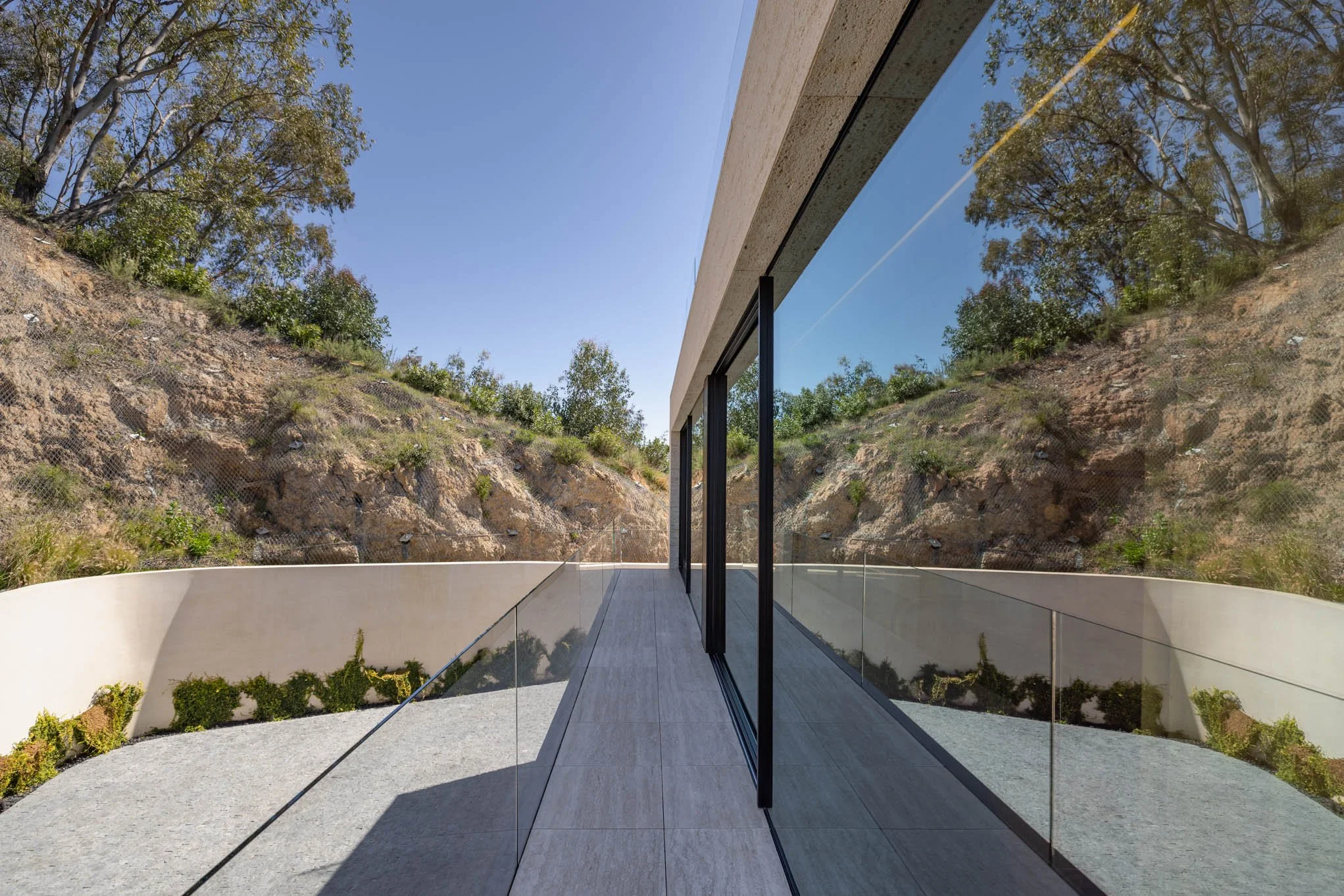 Modern balcony with glass railing and sliding glass doors, overlooking a hillside with trees and rocks under a clear blue sky.