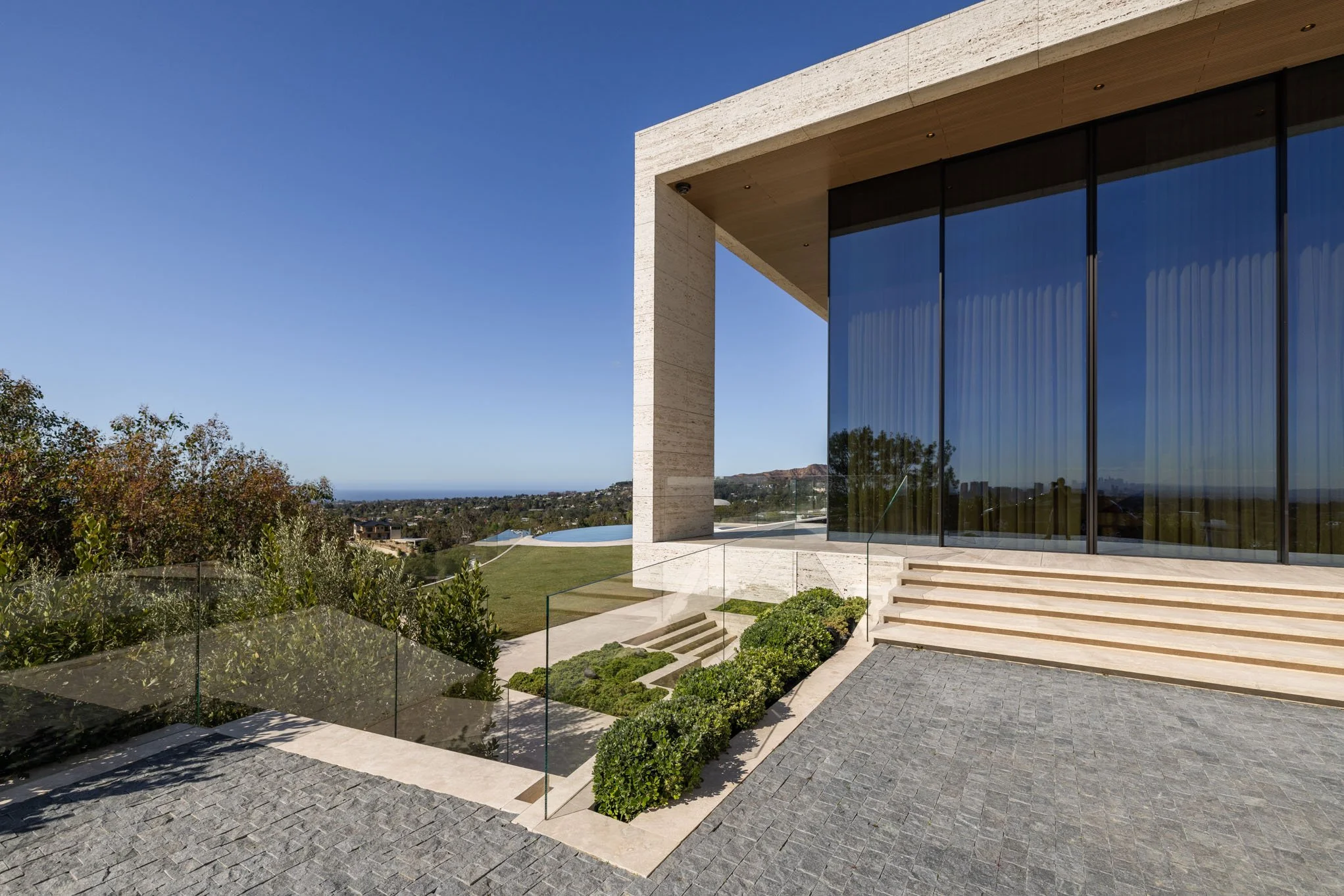 Modern house exterior with large glass doors, stone stairs, and landscaped garden overlooking a grassy area and distant hills under a clear blue sky.