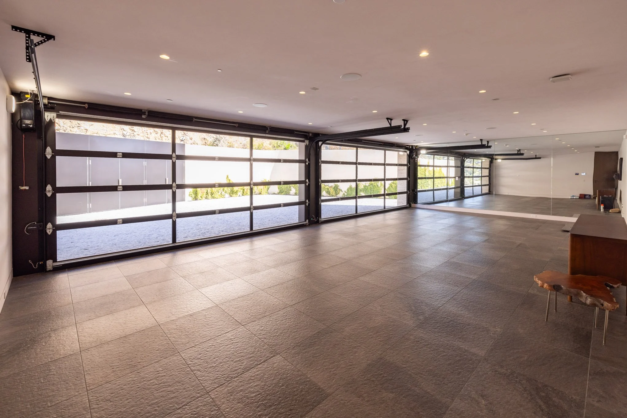 Empty indoor garage with large glass garage doors and a reflection of wall mirror, wooden bench, and side table on the right.