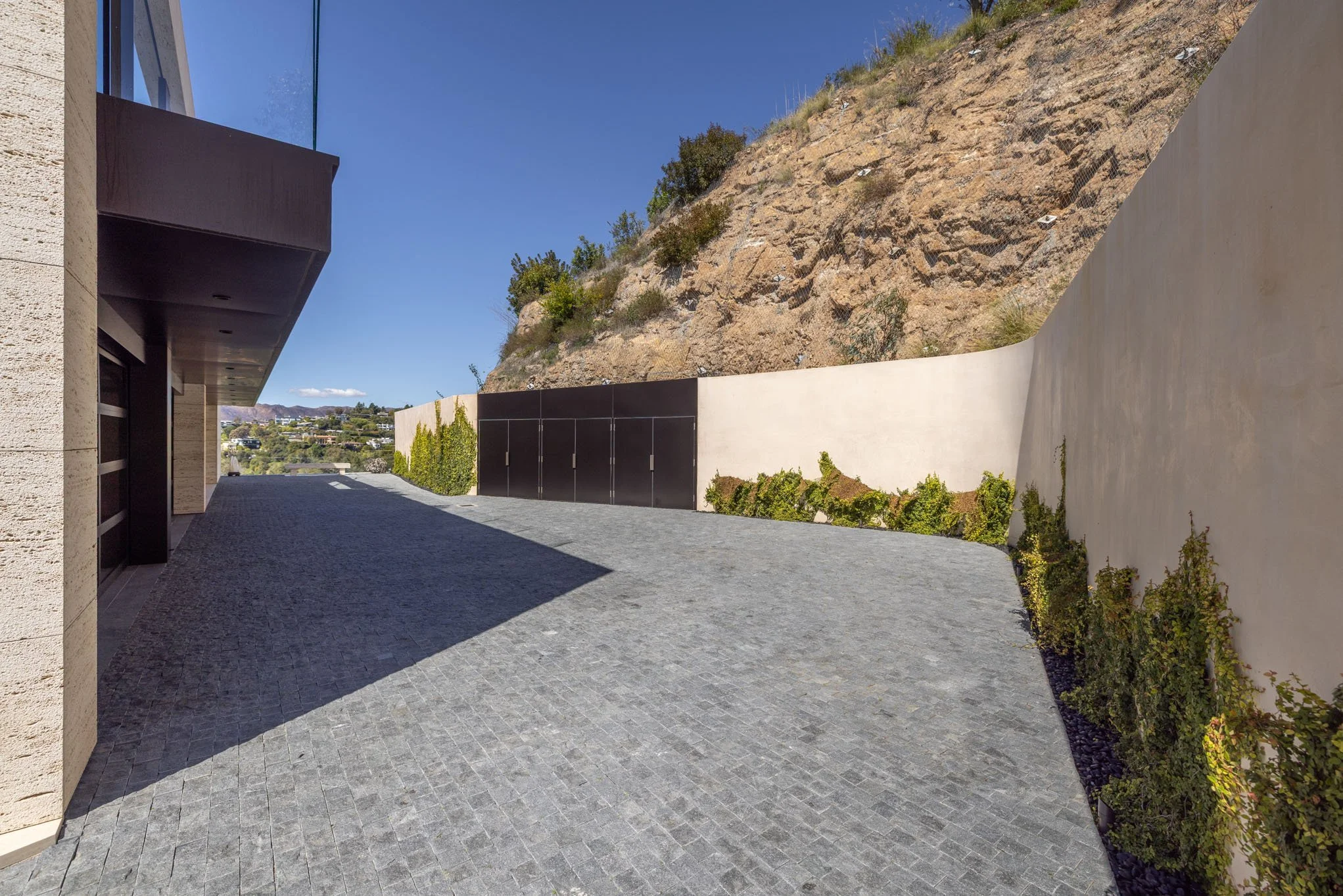 Modern driveway with gray pavers, tall beige wall with green plants, and rocky hill in the background under a blue sky.