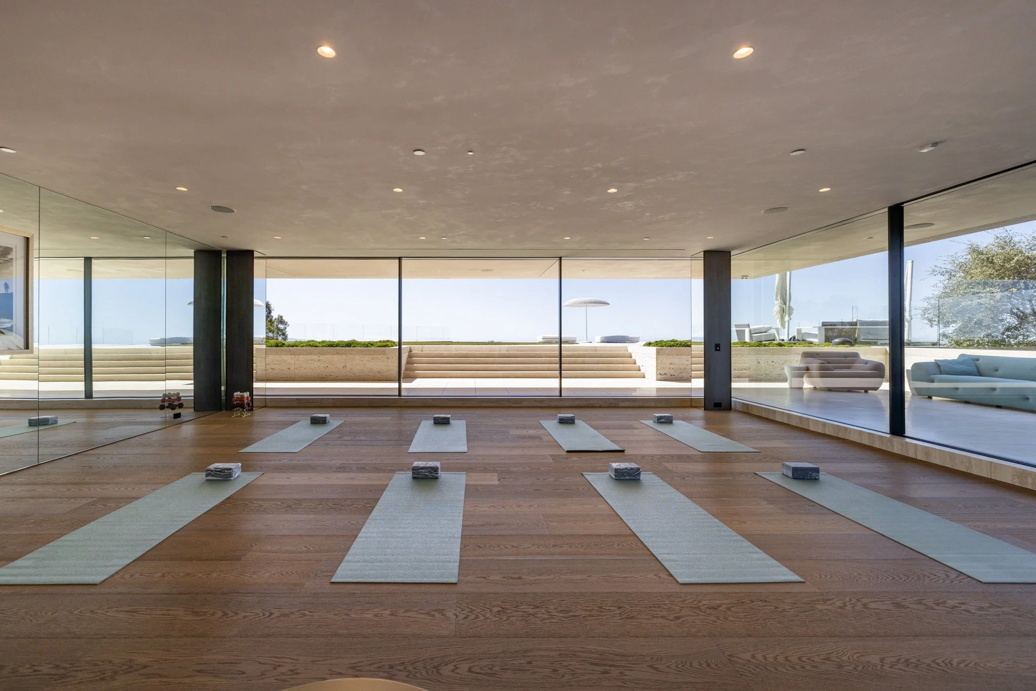 A well-lit indoor yoga studio with wooden floors, six yoga mats with folded towels, and large glass windows viewing an outdoor patio with seating and a clear sky.