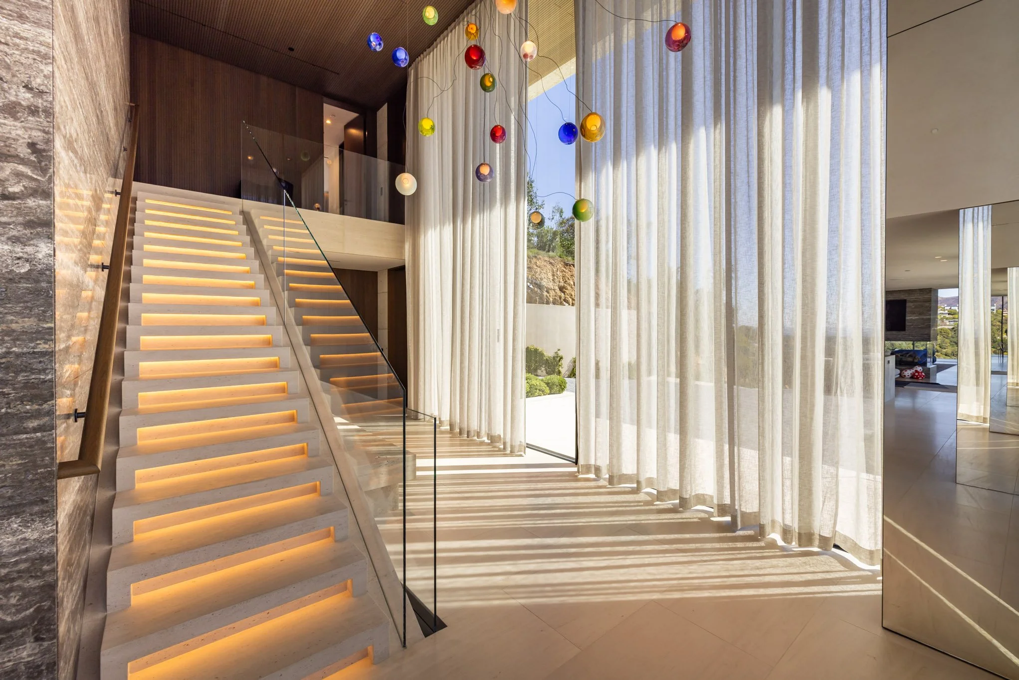Modern interior with marble staircase, floor-to-ceiling curtains, and colorful hanging light orbs.