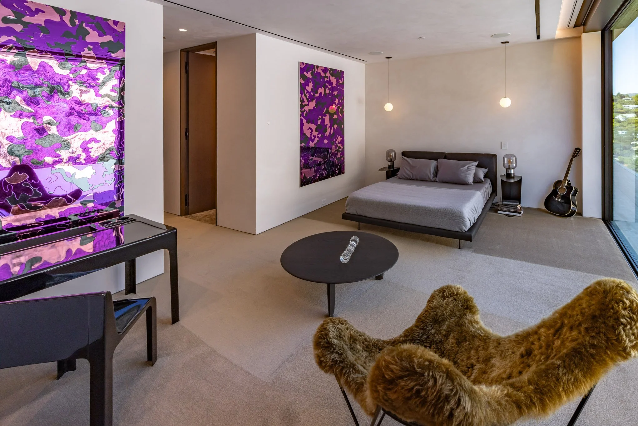 Modern bedroom with a large window, minimalist decor, a bed with grey bedding, two nightstands with lamps, a guitar next to the window, and artwork on the walls. There is a black table with a glass sculpture, a furry lounge chair, and a purple abstra