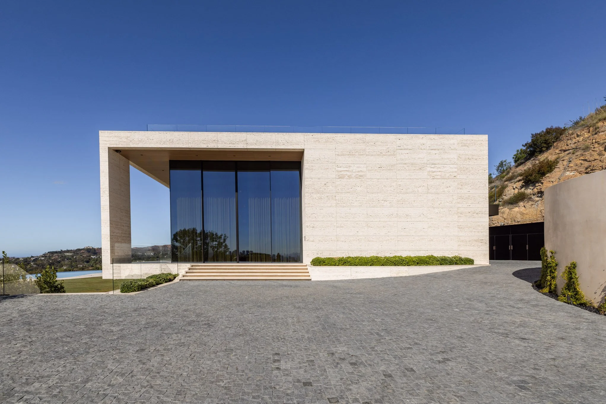 Modern minimalist white building with large glass doors, steps leading up, surrounded by a paved area, with a hillside on the right and a clear blue sky overhead.