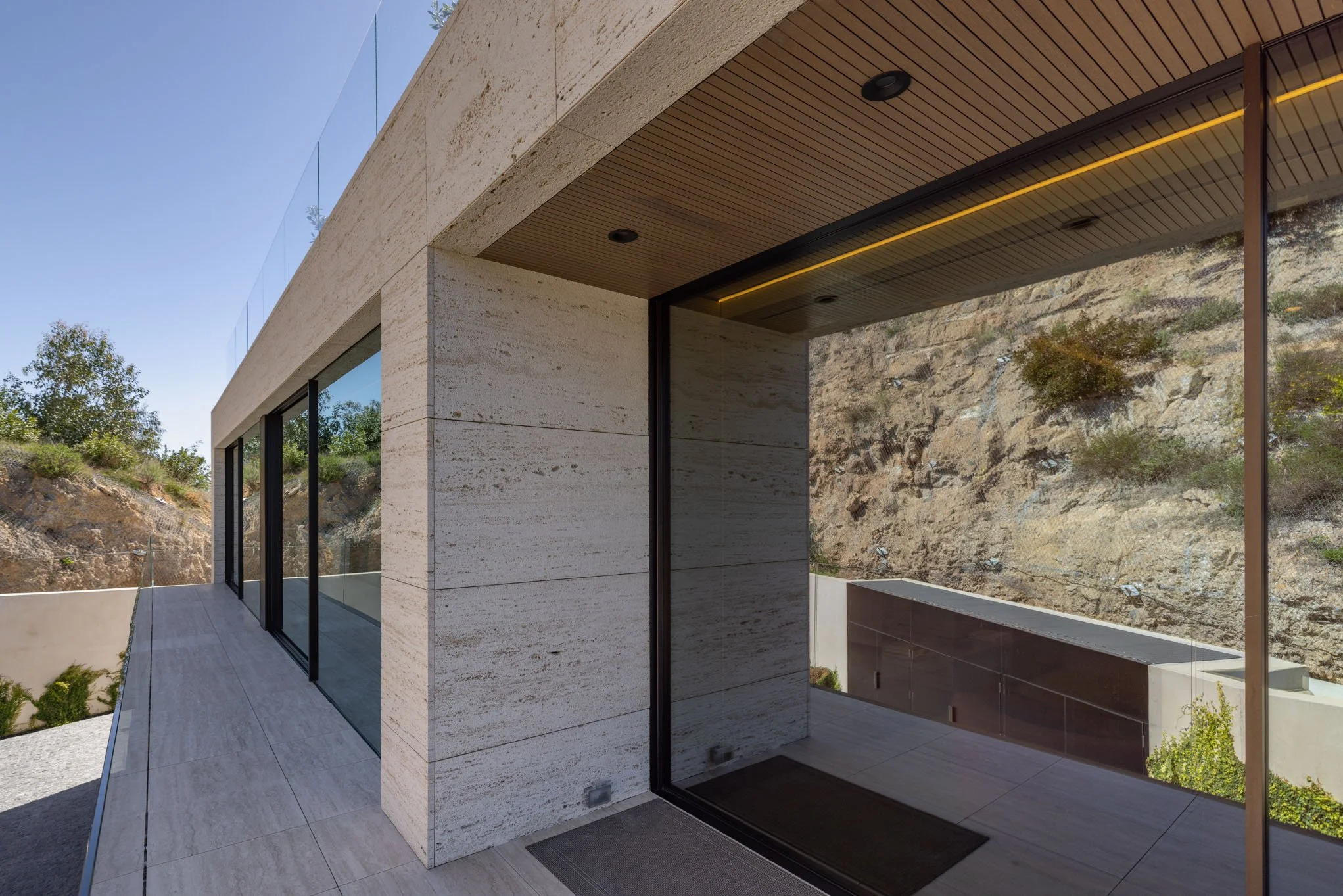 Modern house exterior with large glass windows, stone walls, and a balcony overlooking a rocky hillside and greenery.