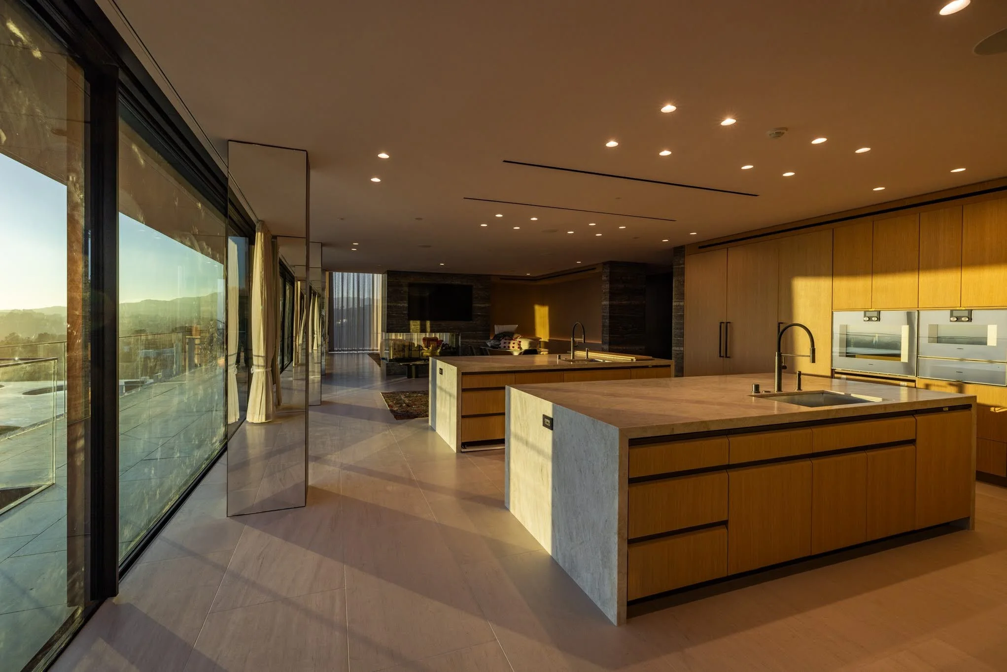 Modern kitchen with large islands, wooden cabinets, and floor-to-ceiling windows showing an outdoor view, illuminated by recessed ceiling lights.