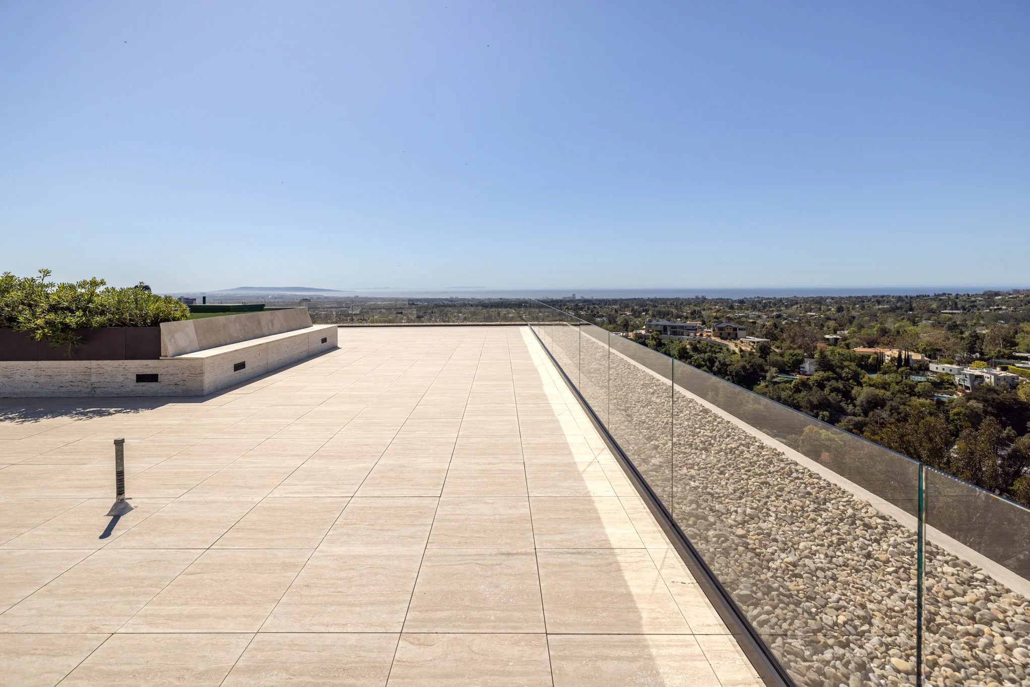 A spacious rooftop terrace with beige tiled flooring, a glass railing on the right, and a small garden area with green plants on the left. The view overlooks a cityscape with trees and buildings and a distant ocean horizon beneath a clear blue sky.