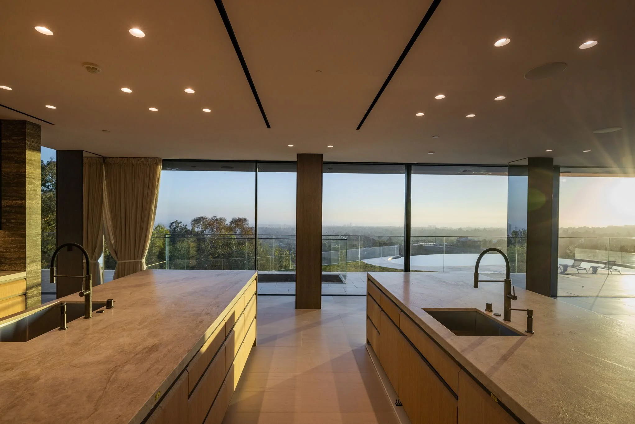 Modern kitchen with two marble islands, stainless steel sinks and faucets, large floor-to-ceiling windows with a scenic view, beige curtains, wood cabinetry, and recessed ceiling lights.