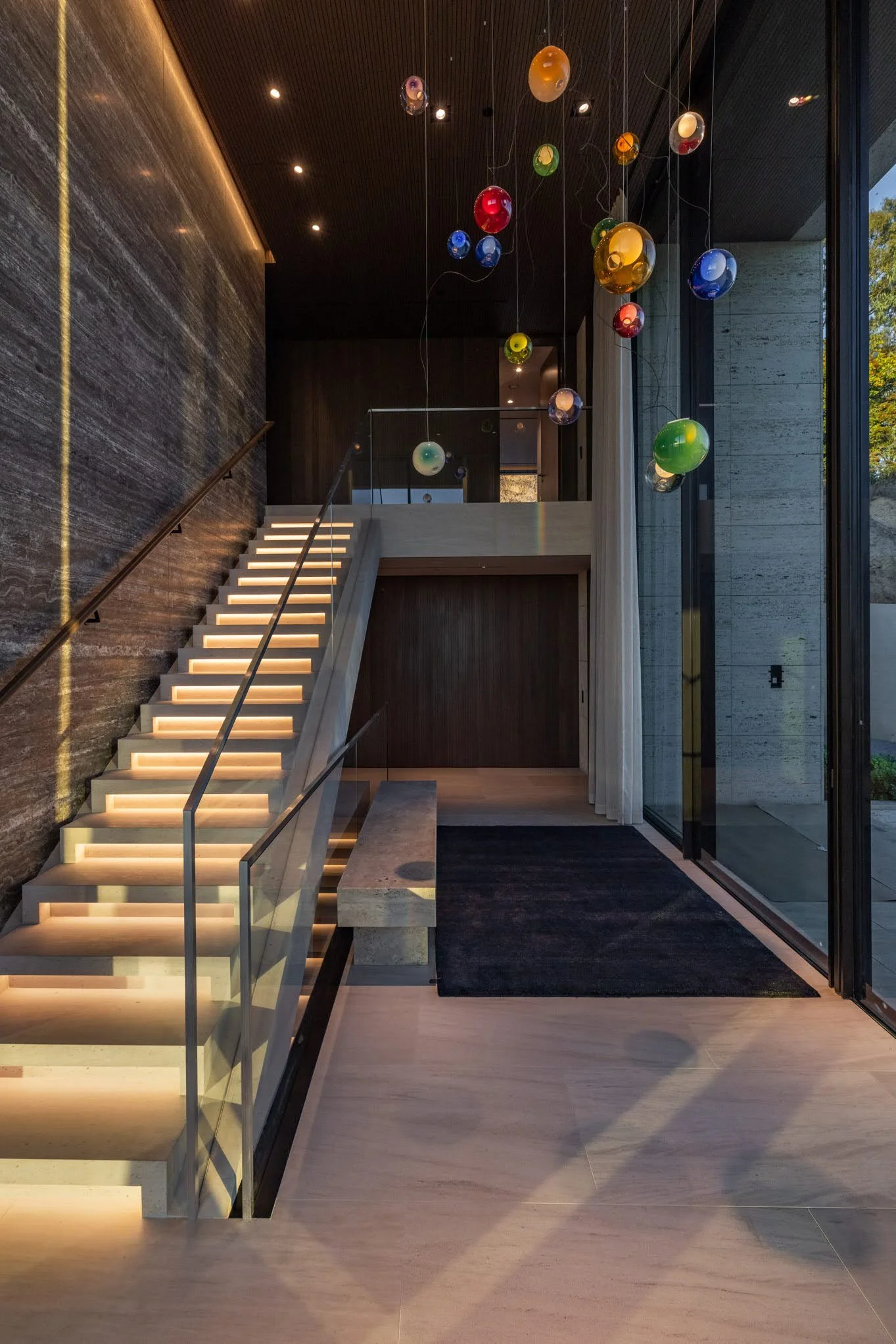Modern interior with illuminated staircase, concrete and wood walls, black rug, glass wall, and colorful hanging glass orb sculptures.