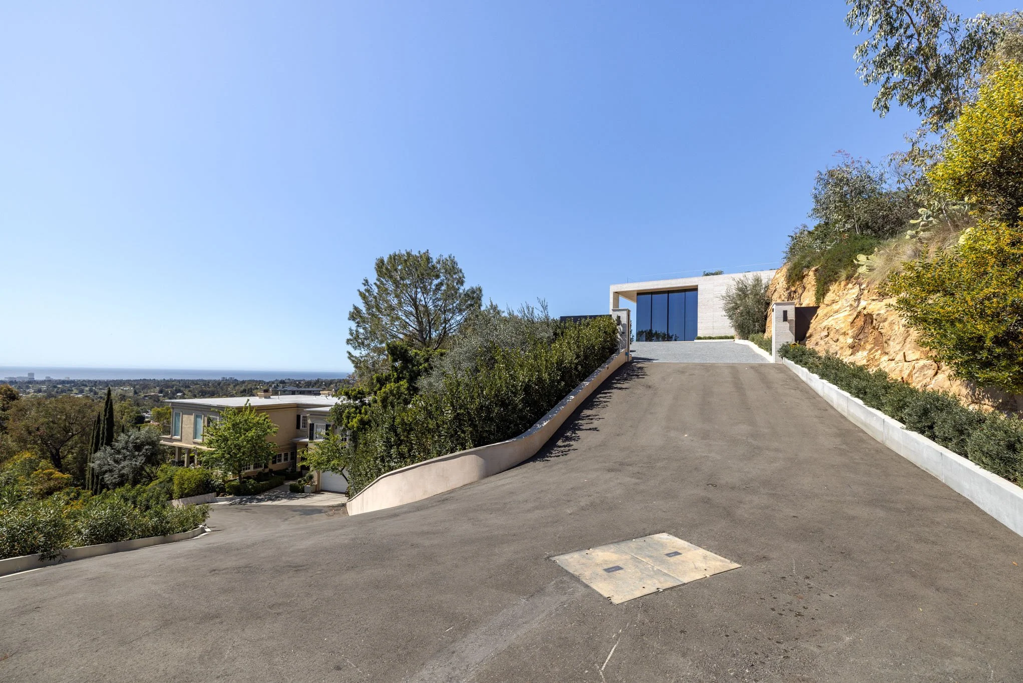 Upside-down view of a modern house on a hill with a steep driveway, garden, trees, shrubs, and city view in the distance under a clear blue sky.
