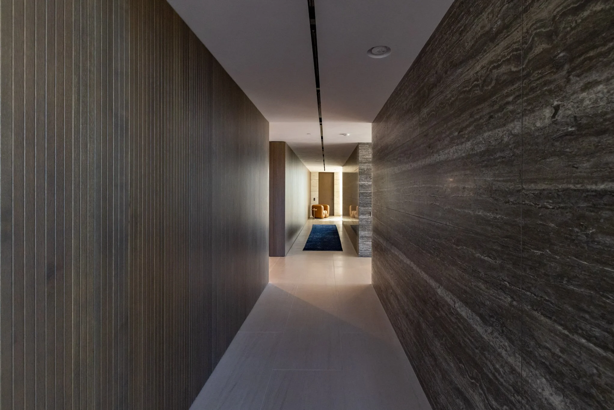 Modern hotel corridor with wood and stone wall designs and a blue rug, leading to a sitting area at the end.