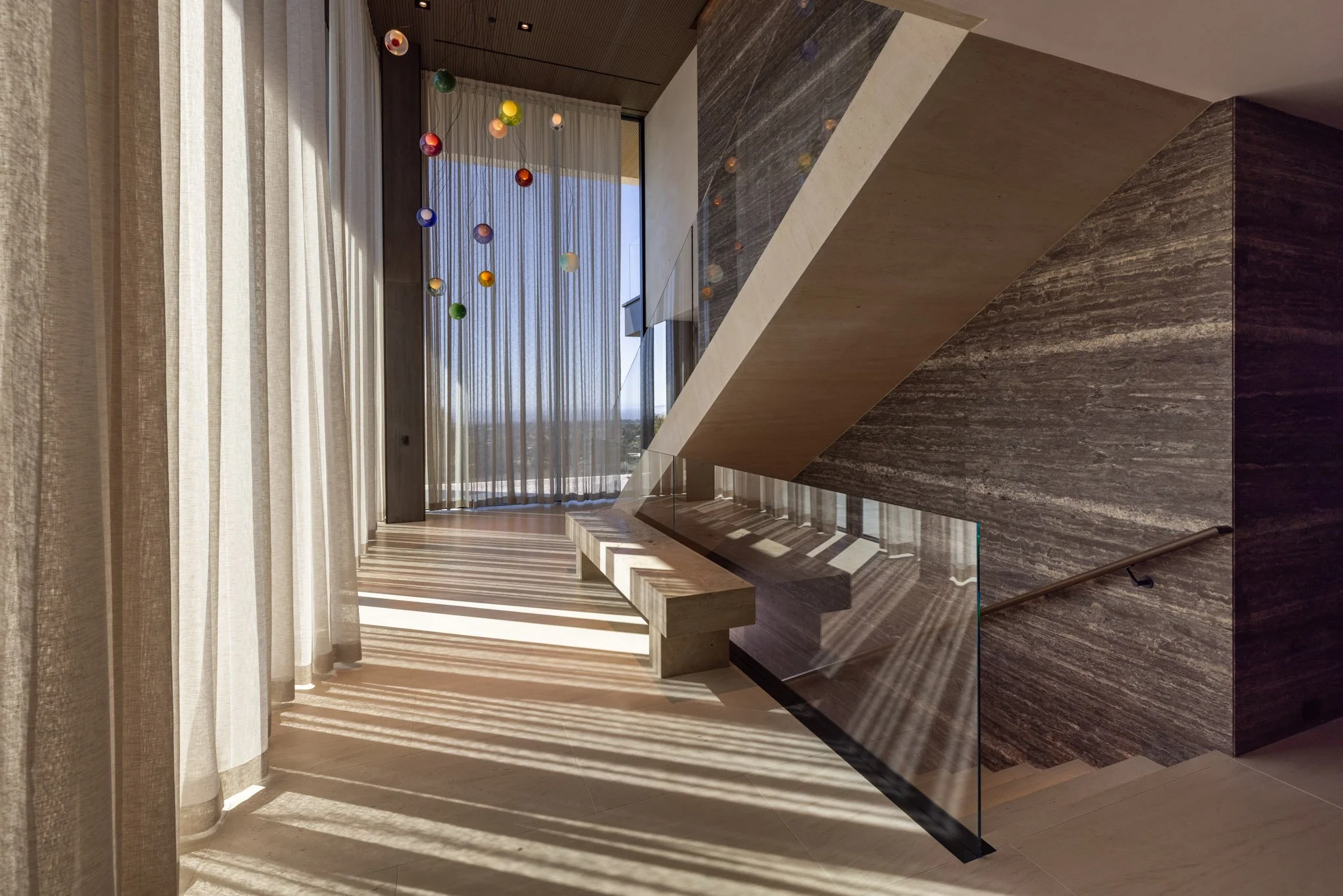 Modern interior staircase with marble wall, glass balustrade, wooden steps, large windows with sheer curtains, and a colorful mobile hanging from the ceiling.