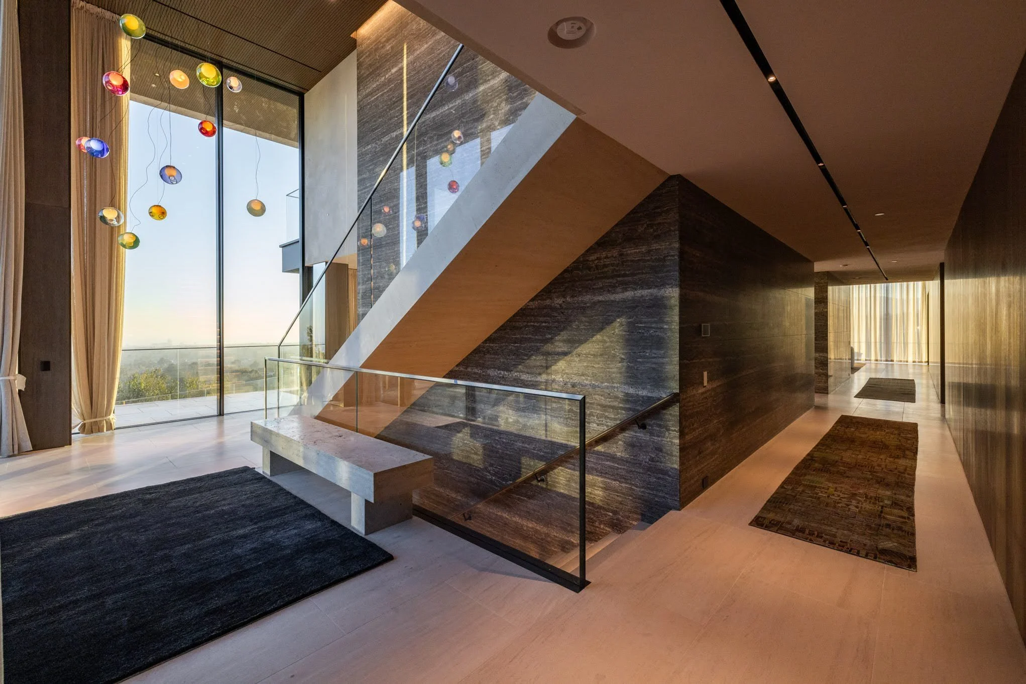 Modern interior with a staircase, large floor-to-ceiling window with curtains, colorful hanging glass orbs, dark textured walls, and light-colored flooring with two rugs.