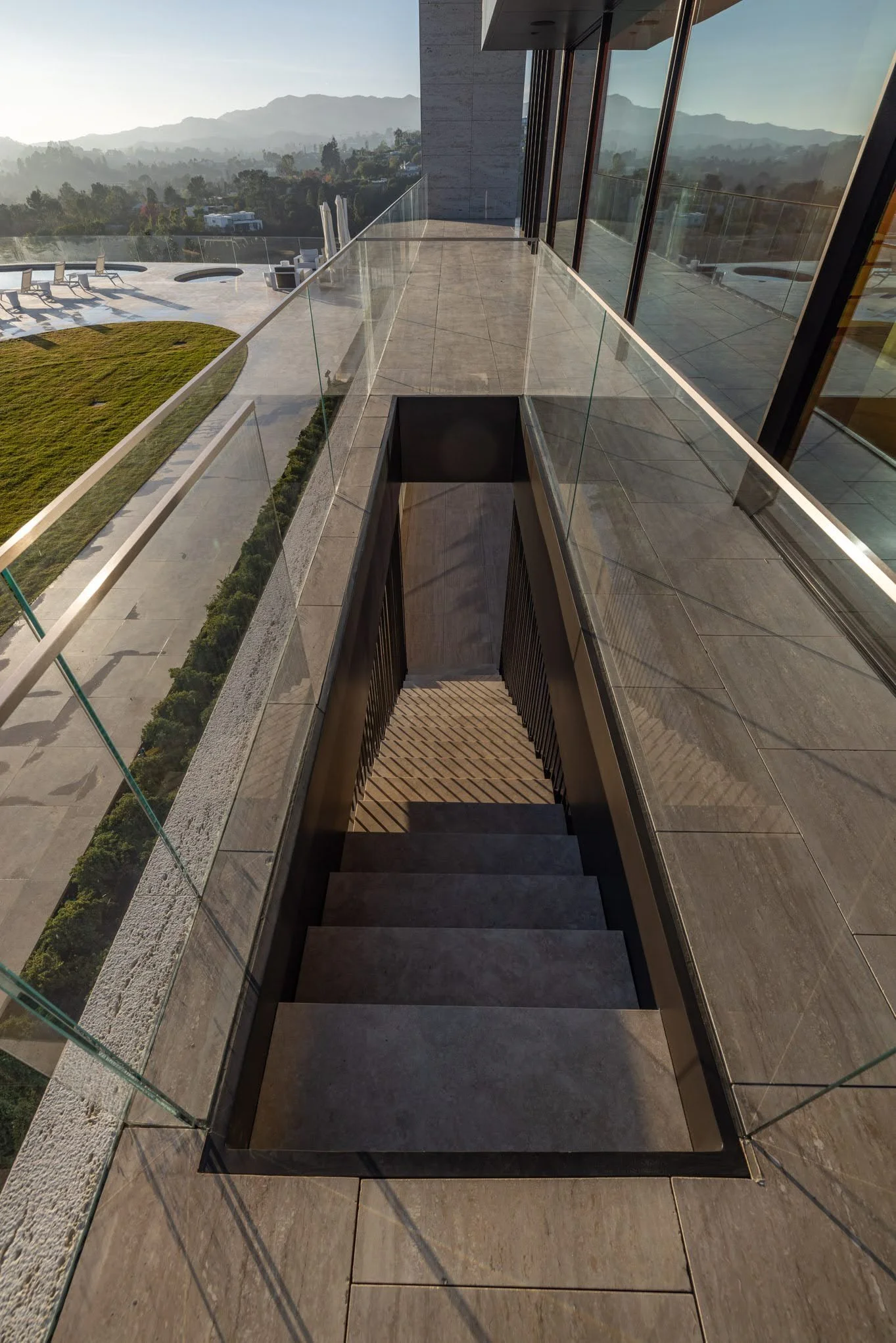 Outdoor balcony with glass railing overlooking a scenic landscape with hills and trees, stairs leading down from the balcony, and a modern building with large windows.