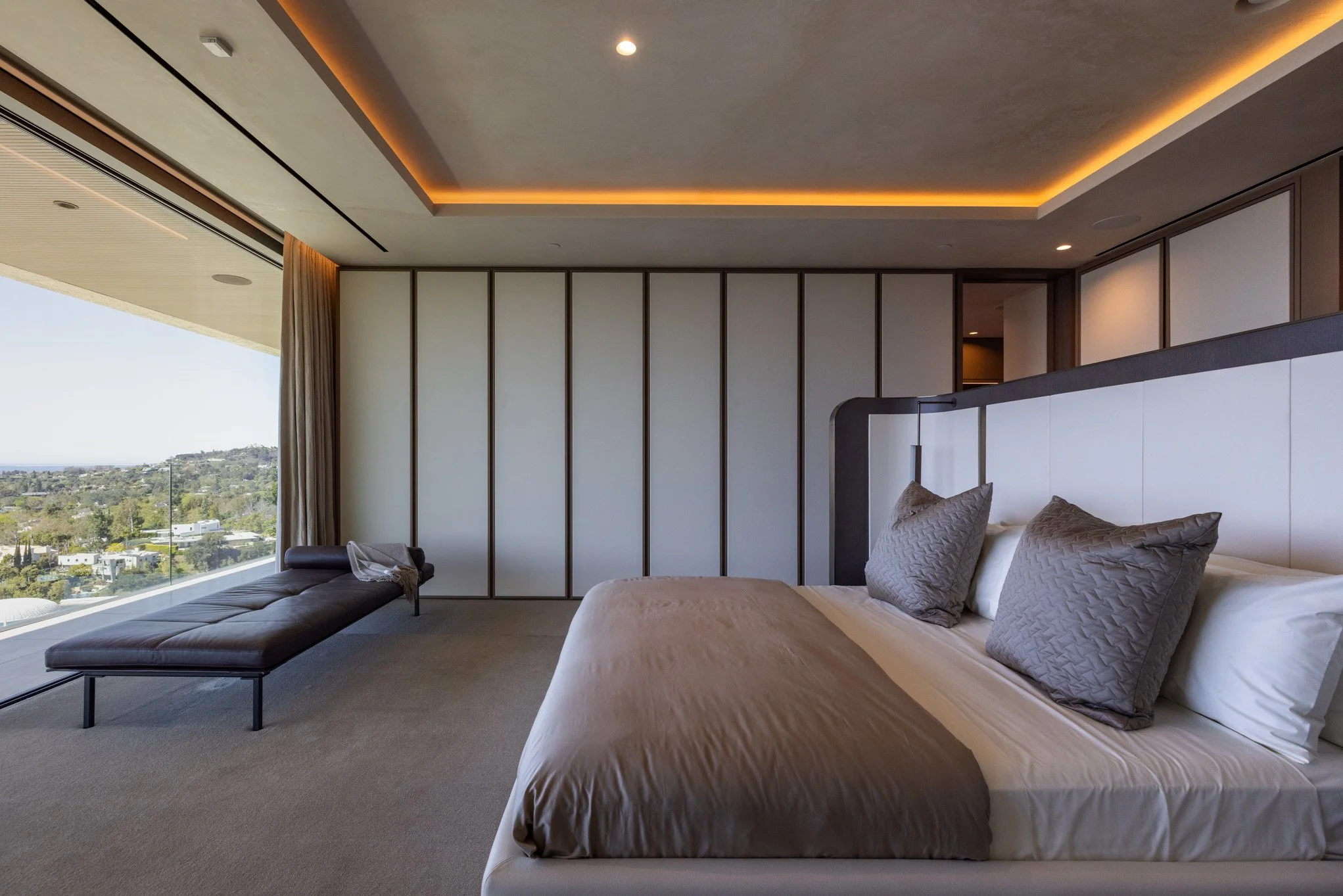 Modern hotel bedroom with a large bed, two gray pillows, and a white headboard, next to a glass wall overlooking a city view, with a bench and minimalist décor.
