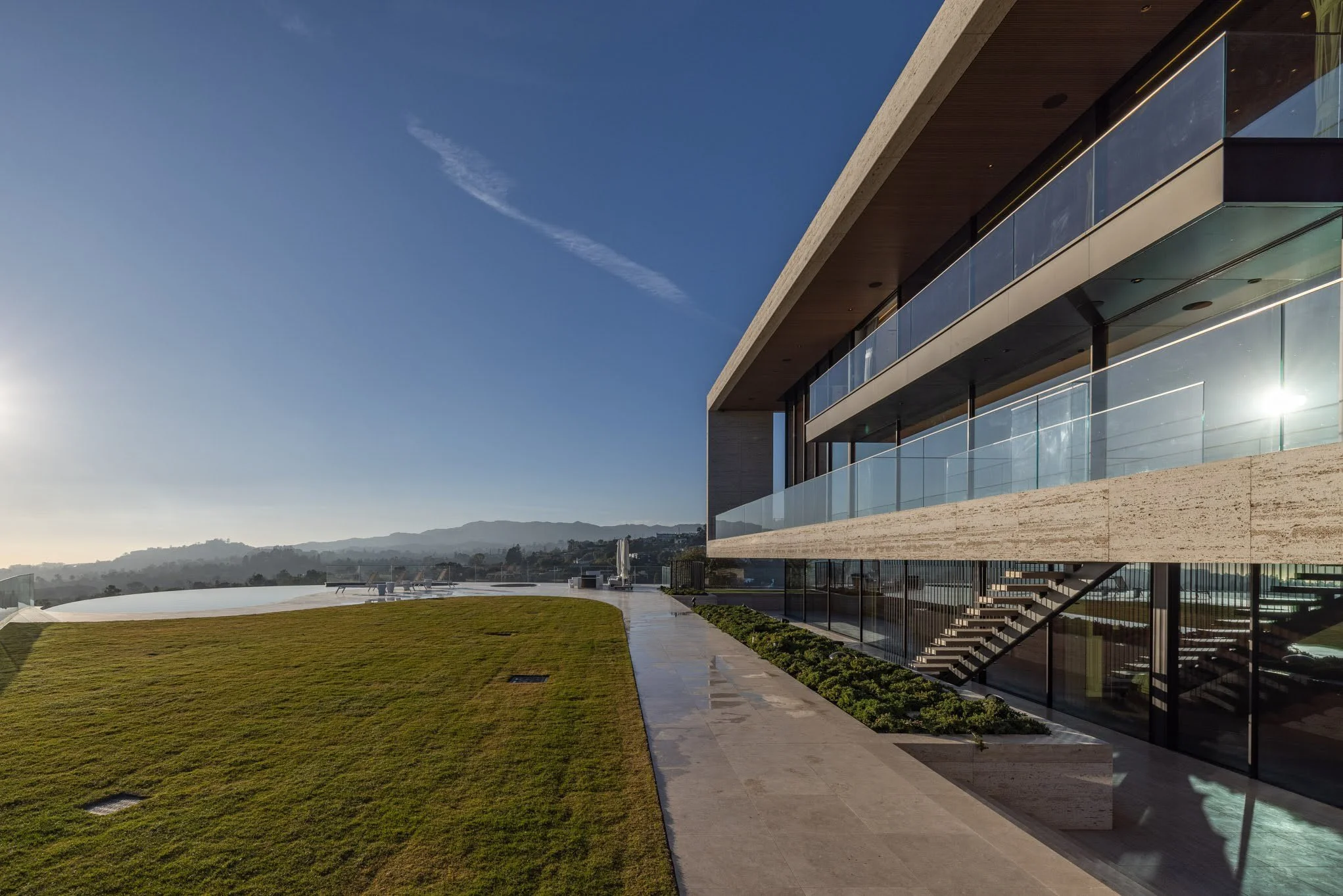 Modern building with large glass windows and balconies, a paved walkway, a grassy area, and a mountain range in the distance under a clear sky with some clouds.