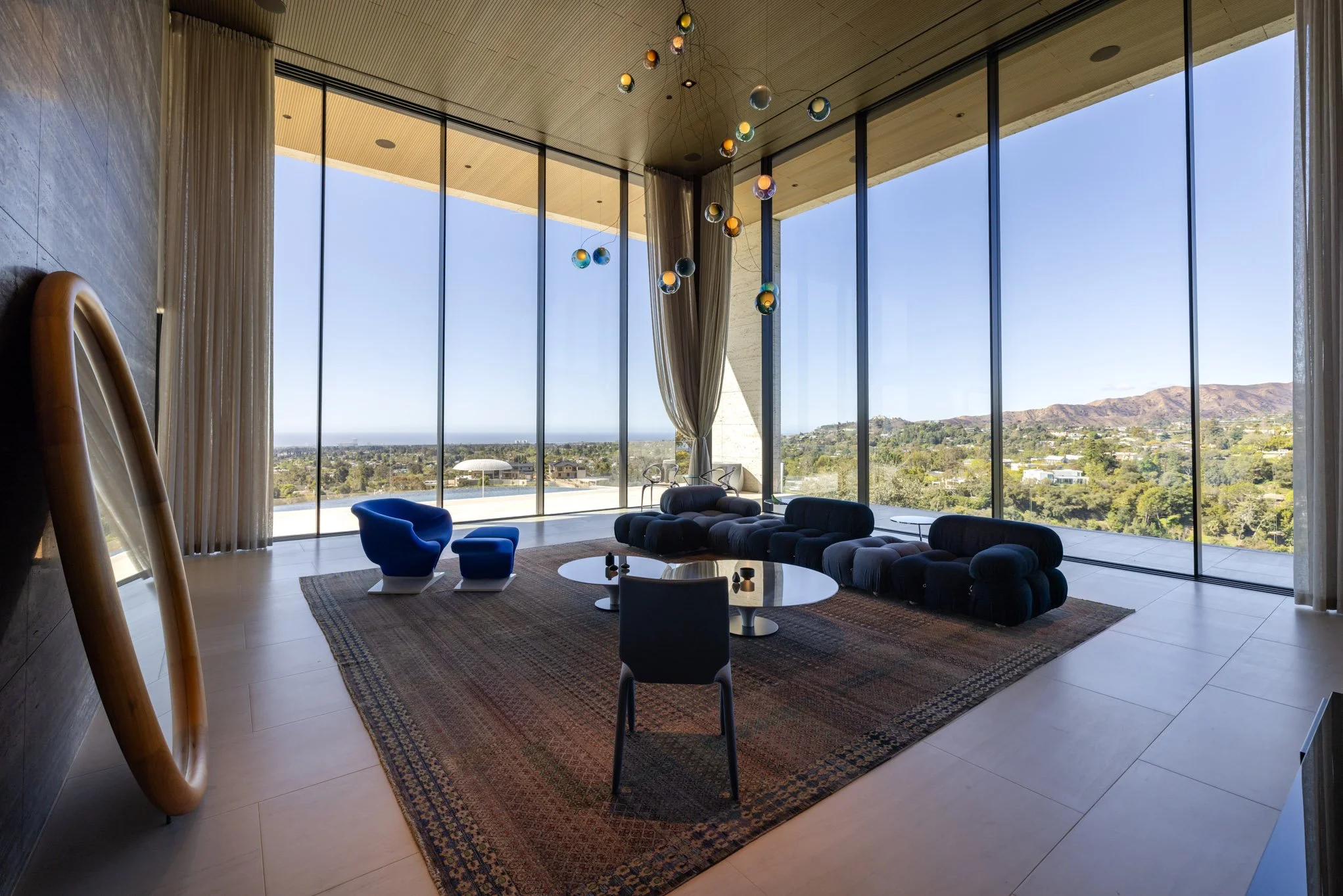 Modern living room with a large floor-to-ceiling glass window view of hills and sky, featuring contemporary furniture including a curved black sofa, a blue lounge chair with matching ottoman, a round glass coffee table, and a black chair. A colorful 