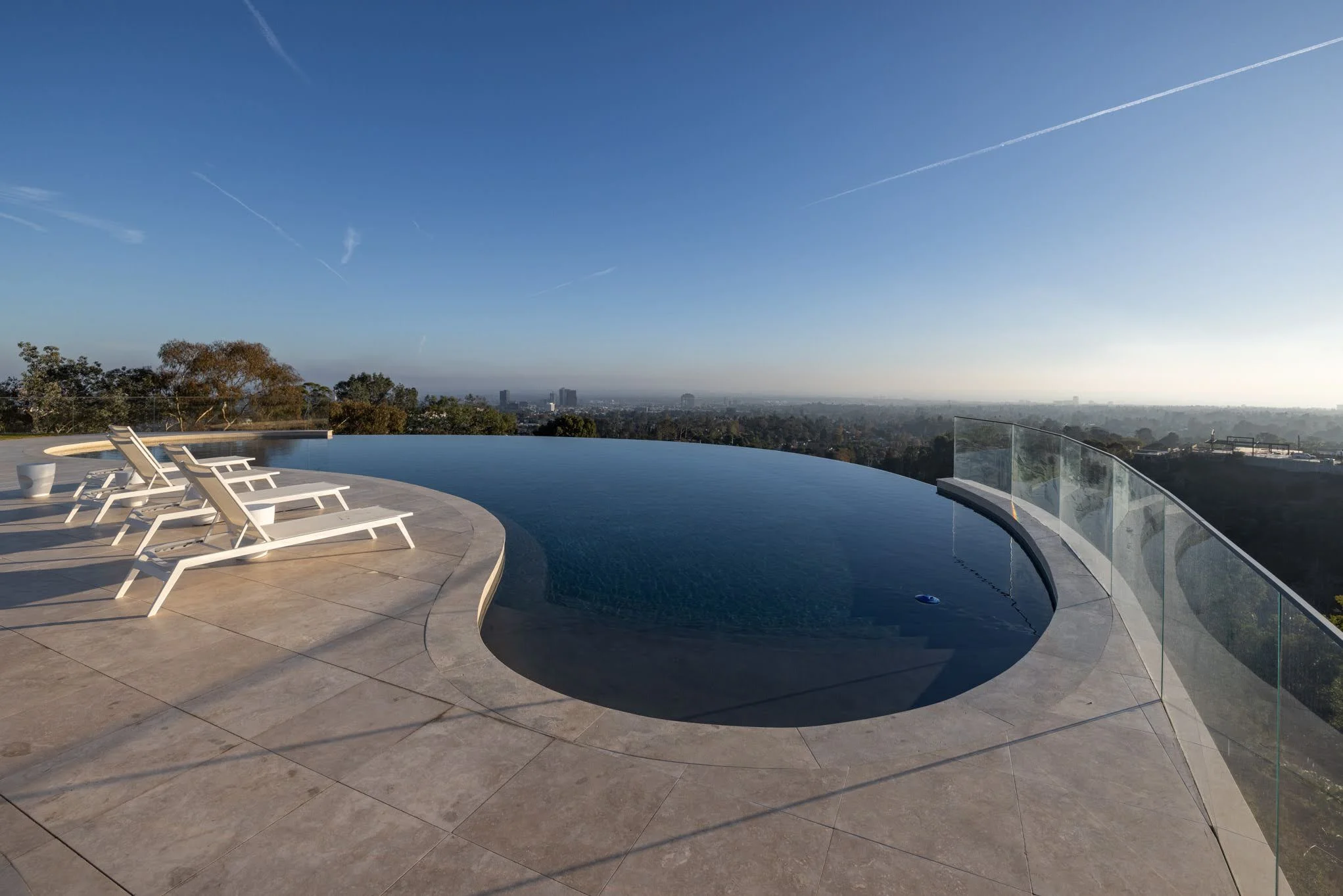 Infinity pool with white lounge chairs and a glass railing overlooking a city skyline on a clear day.