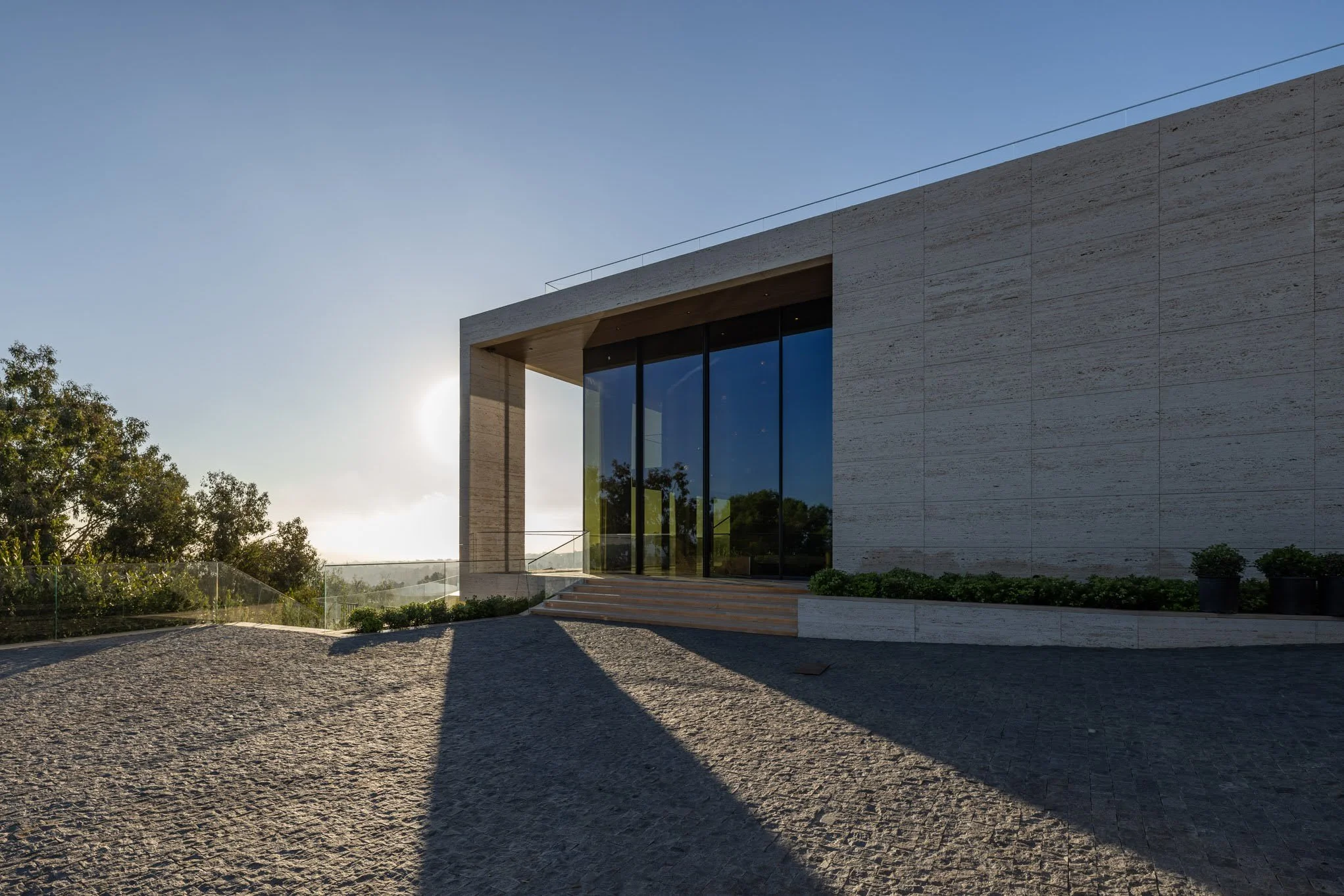 Modern building with large glass doors, stone exterior, and steps leading up to the entrance, with shadow cast on cobblestone paving and trees visible in the background during sunset.