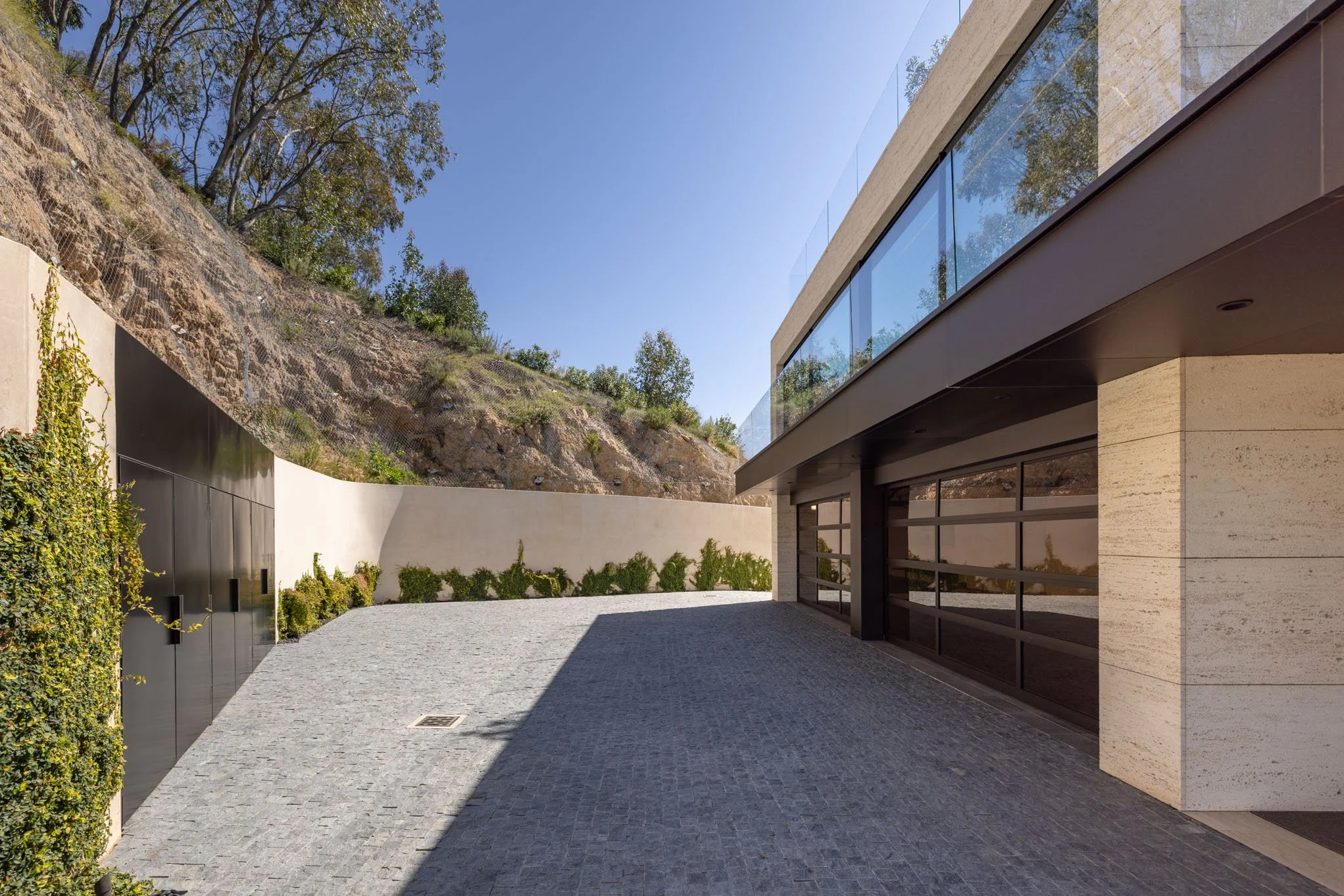 Modern house with large glass windows and a driveway, with a hillside and greenery in the background.