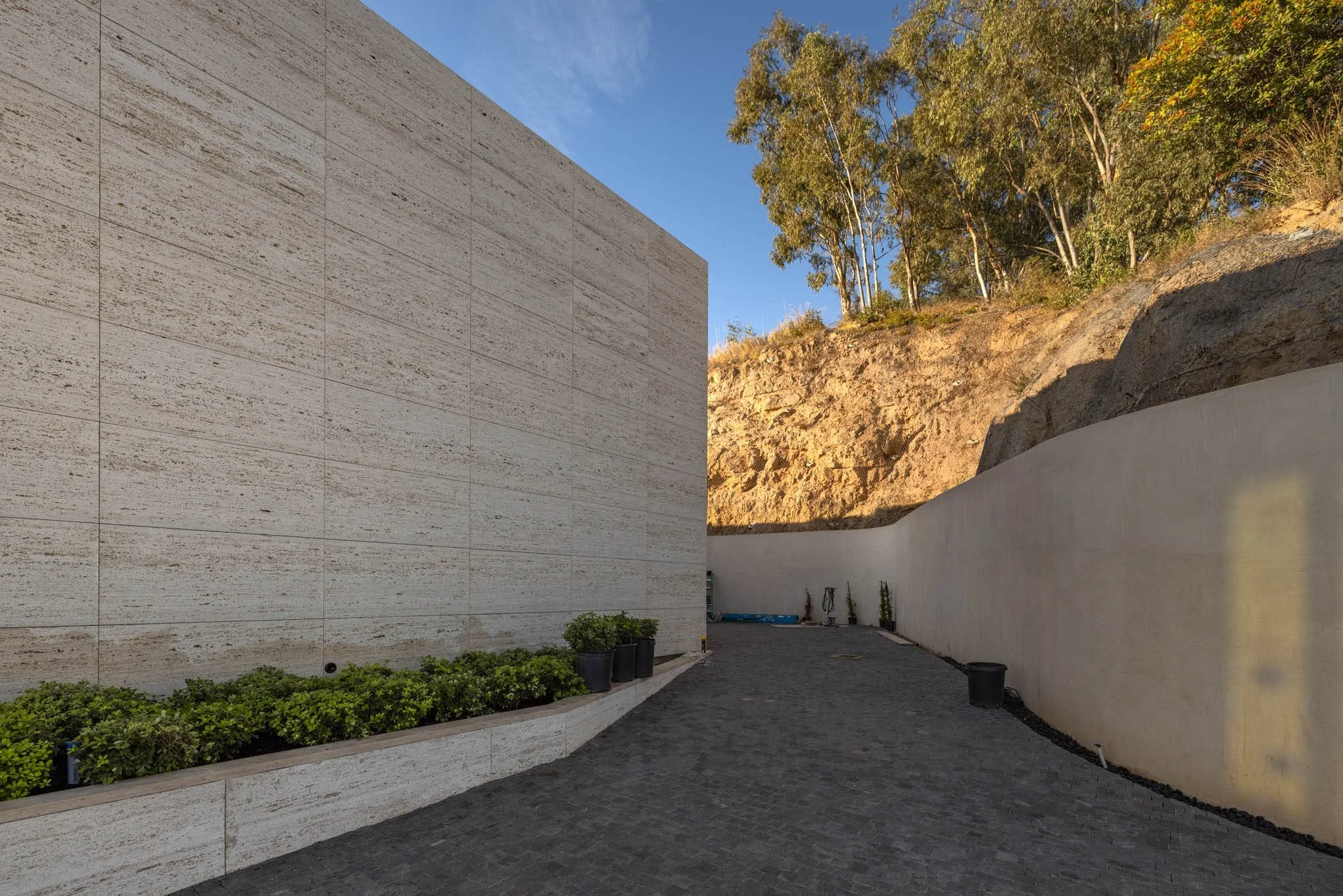 A narrow outdoor pathway between a large beige stone wall on the left and a curved white wall on the right, with potted plants along the base of the beige wall and a hill with trees in the background under a clear blue sky.