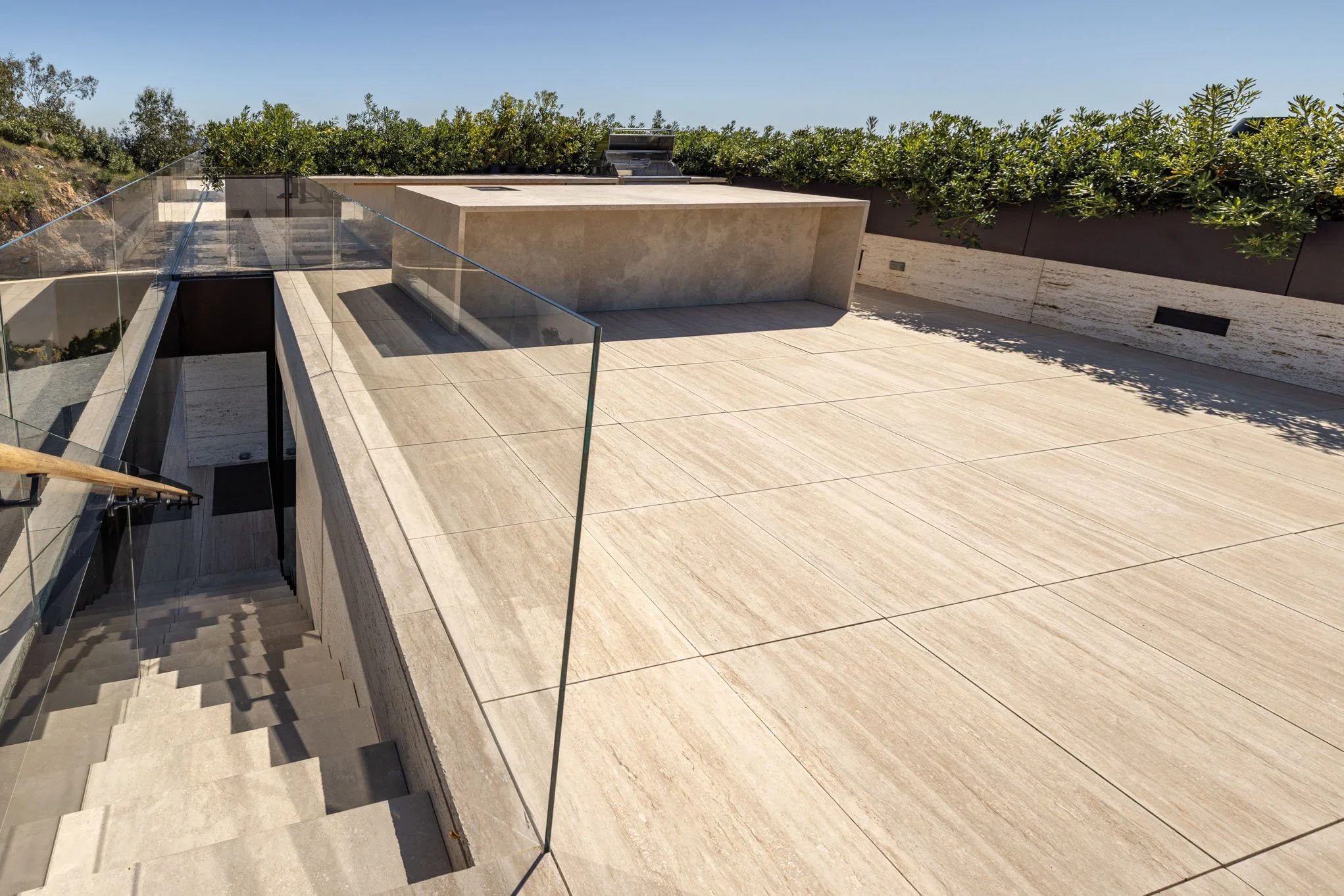 Modern outdoor terrace with beige tiled flooring, glass railing, and greenery along the wall, overlooking a staircase leading down.