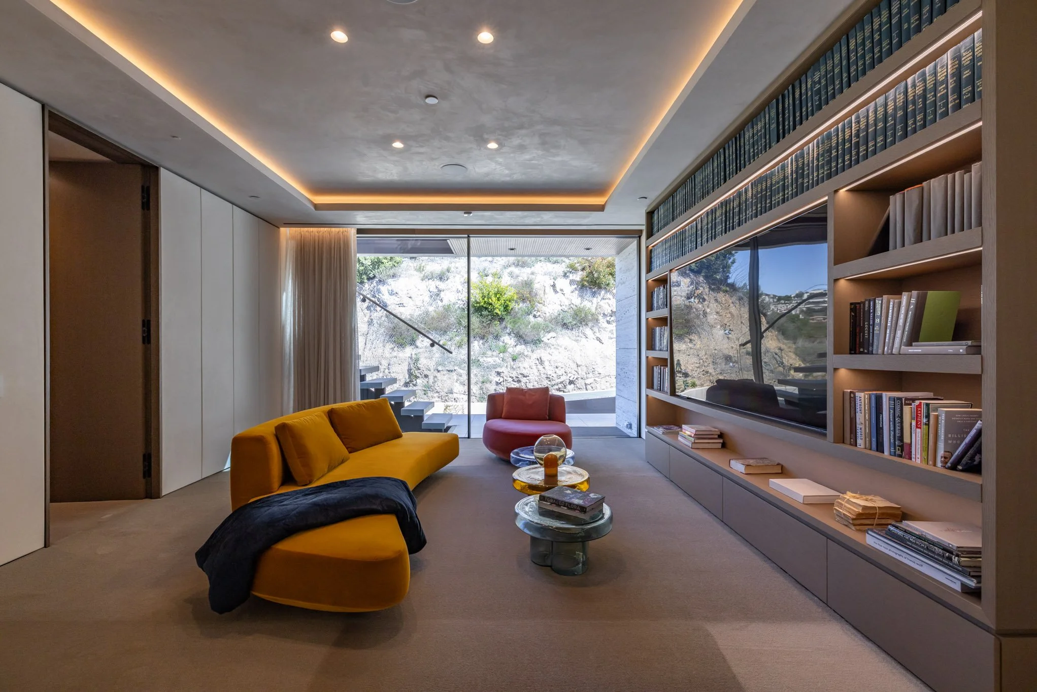 Modern living room with a yellow sofa, pink armchair, marble coffee tables, large built-in bookshelf, and a window overlooking a hillside.