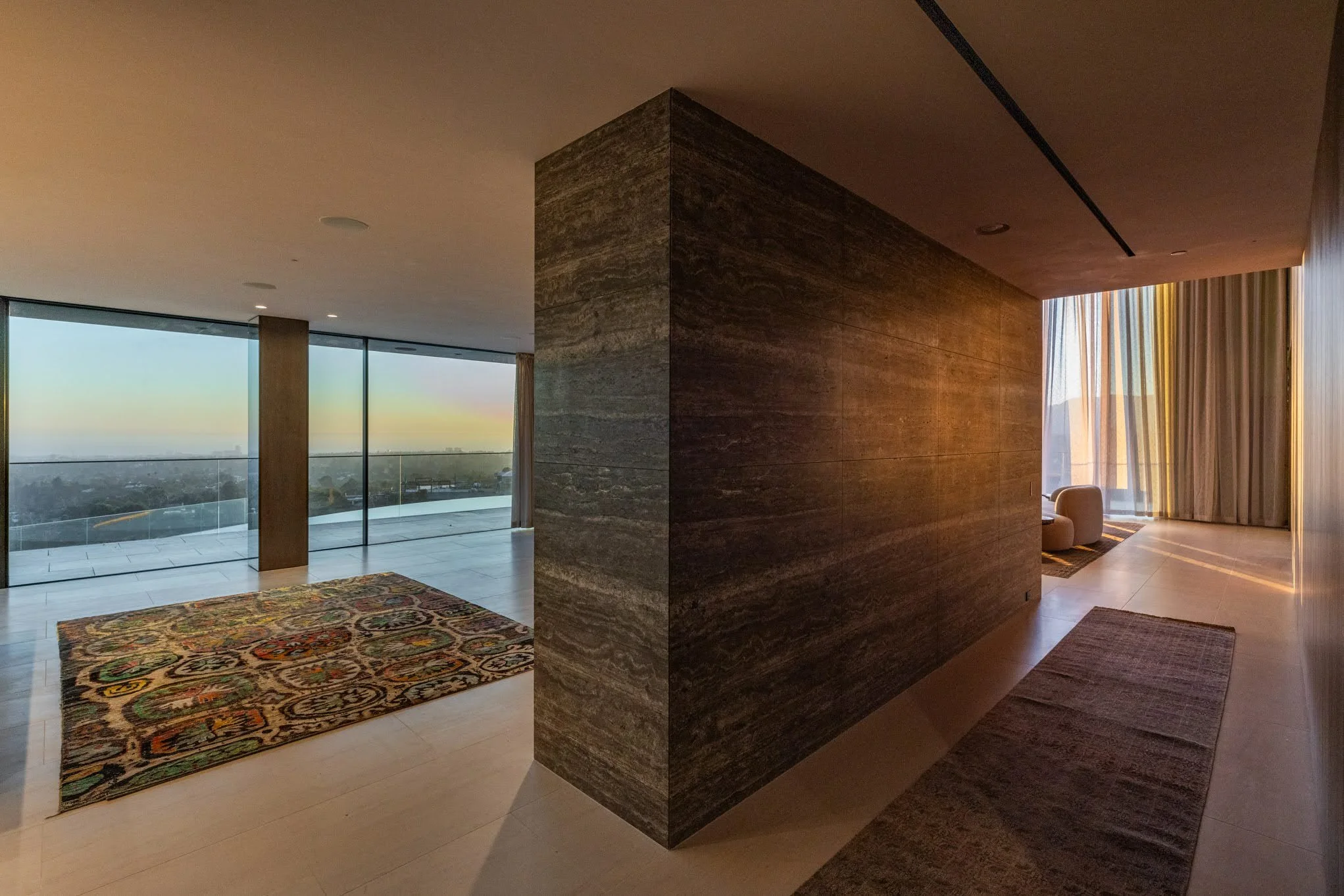 Modern living space with large windows, a colorful area rug on light-colored floor, a dark wall or partition, and a seating area with curtains and sunlight