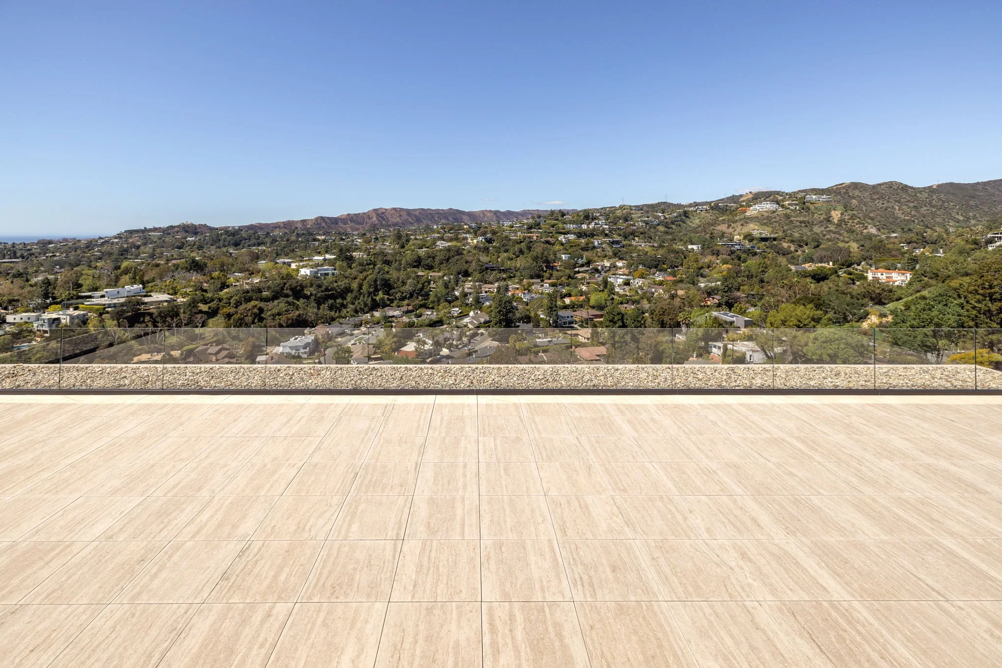 A rooftop terrace with beige tiles and a glass railing overlooking a residential area and hills in the distance under a clear blue sky.