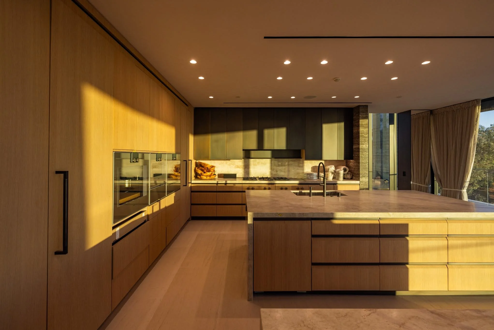 Modern kitchen with wooden cabinets, marble countertops, and a large island with a sink, illuminated by sunlight through large windows with cream curtains.