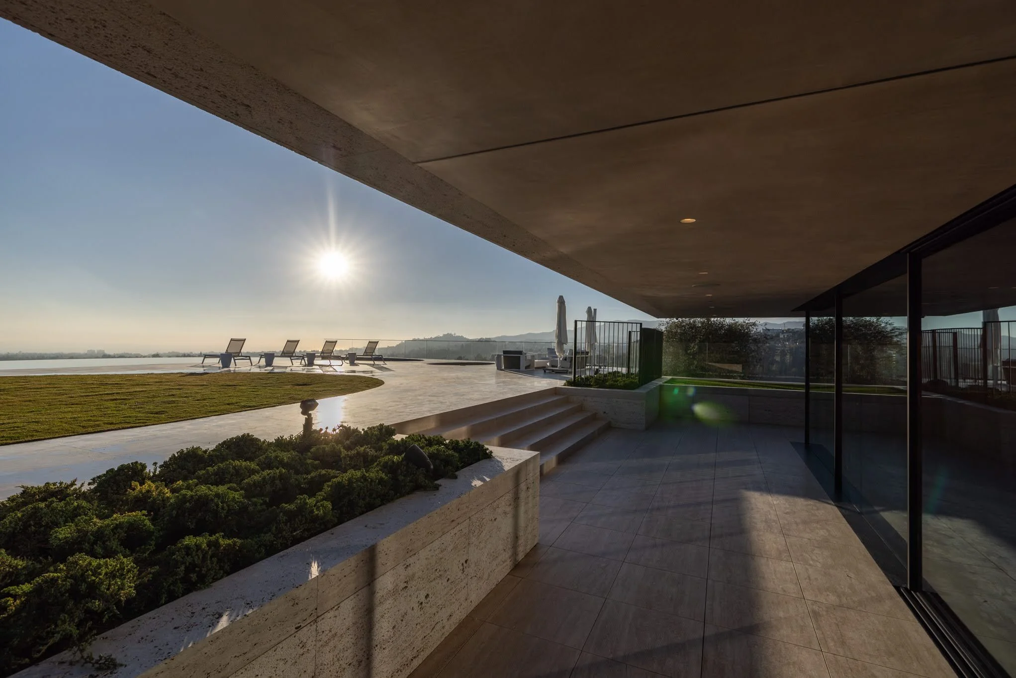 Outdoor terrace with lounge chairs, a lawn, and scenic view of hills and water, under a clear sky with the sun