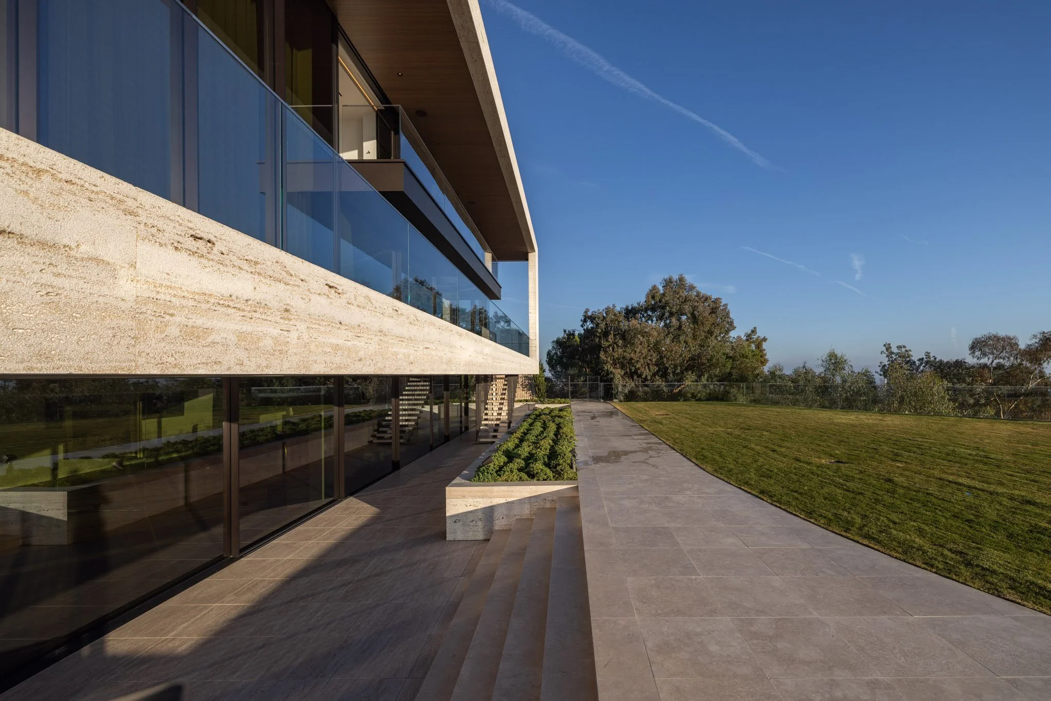 Modern building with large glass windows, a stone exterior, and a landscaped outdoor patio area with grass and trees in the background under a clear blue sky.
