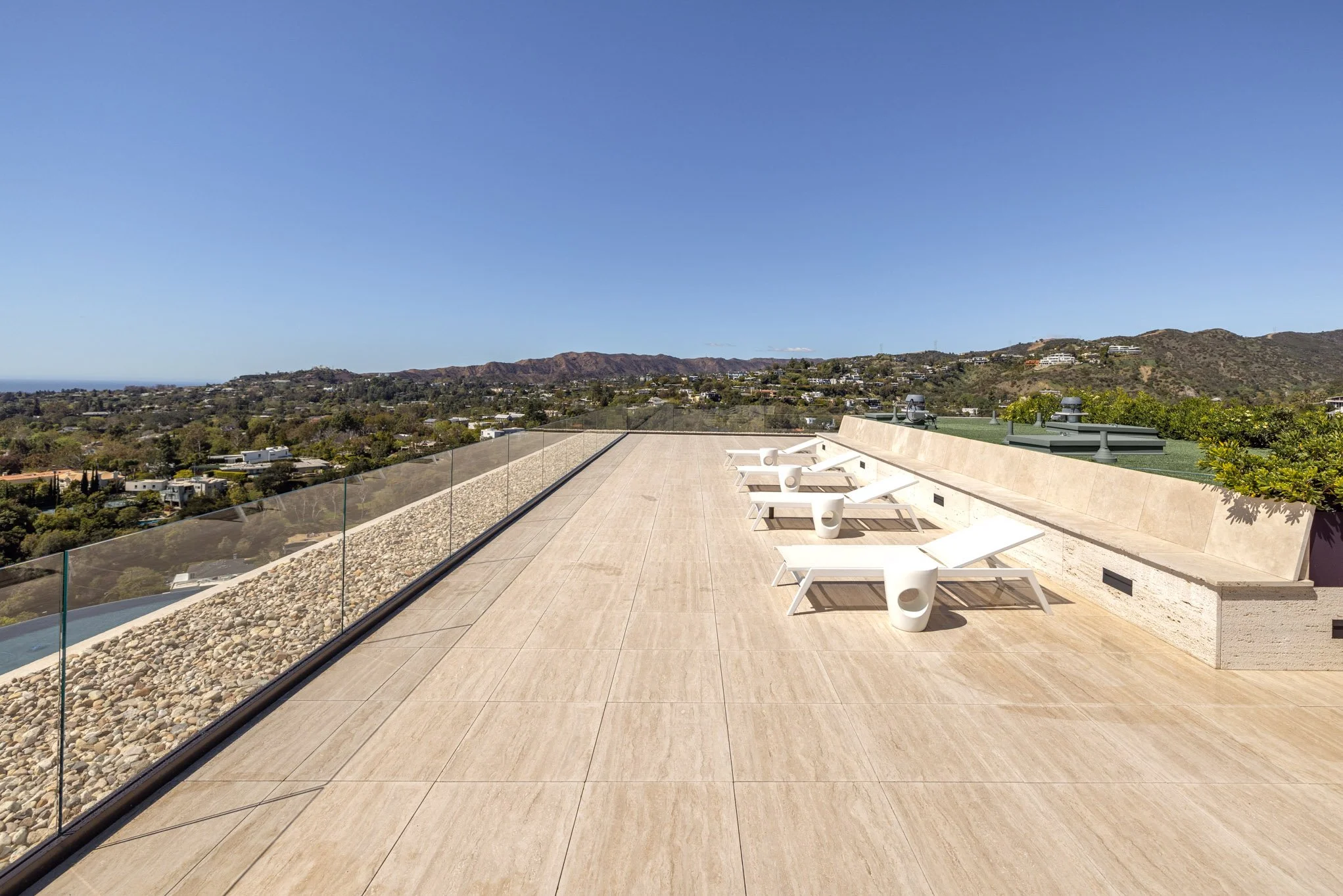 Rooftop terrace with a row of white lounge chairs, small white side tables, glass railing, and a view of distant hills under a clear blue sky.