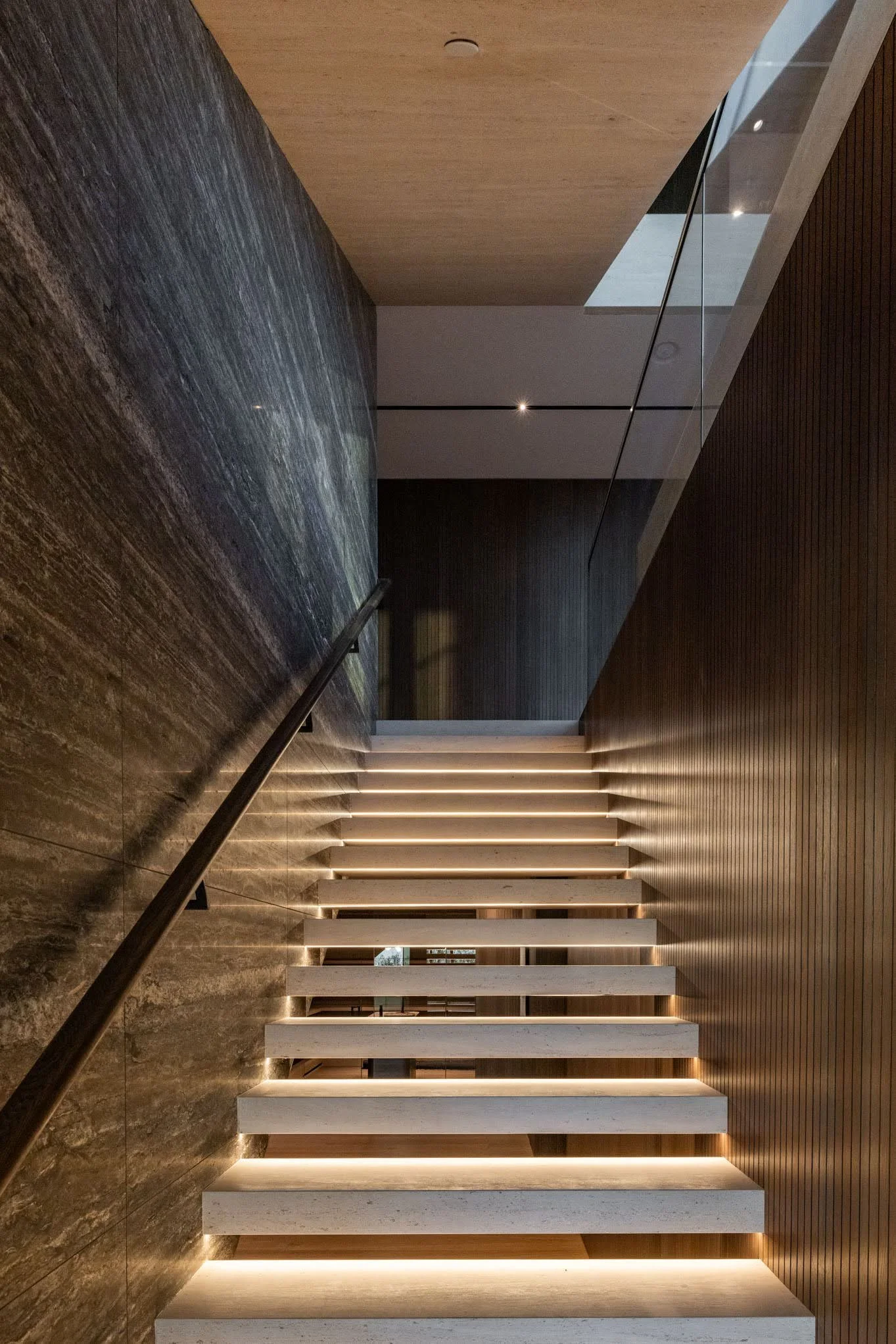 Modern staircase with horizontal white steps and integrated lighting, enclosed by dark and light wood wall panels and a glass railing.