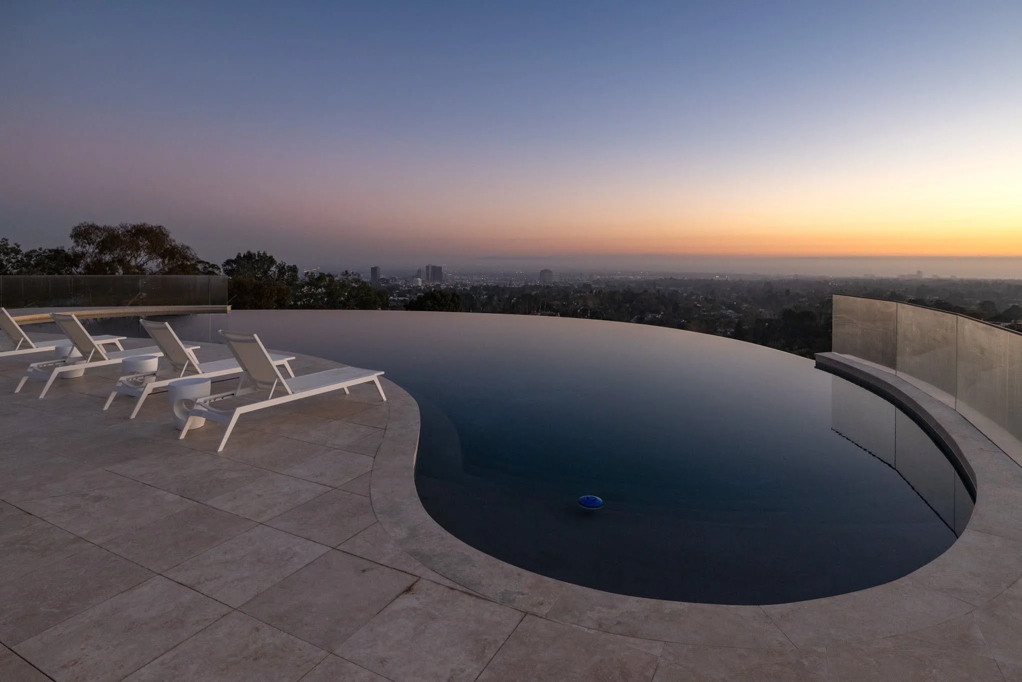Infinity swimming pool on a rooftop at sunset with lounge chairs nearby, overlooking a city skyline.
