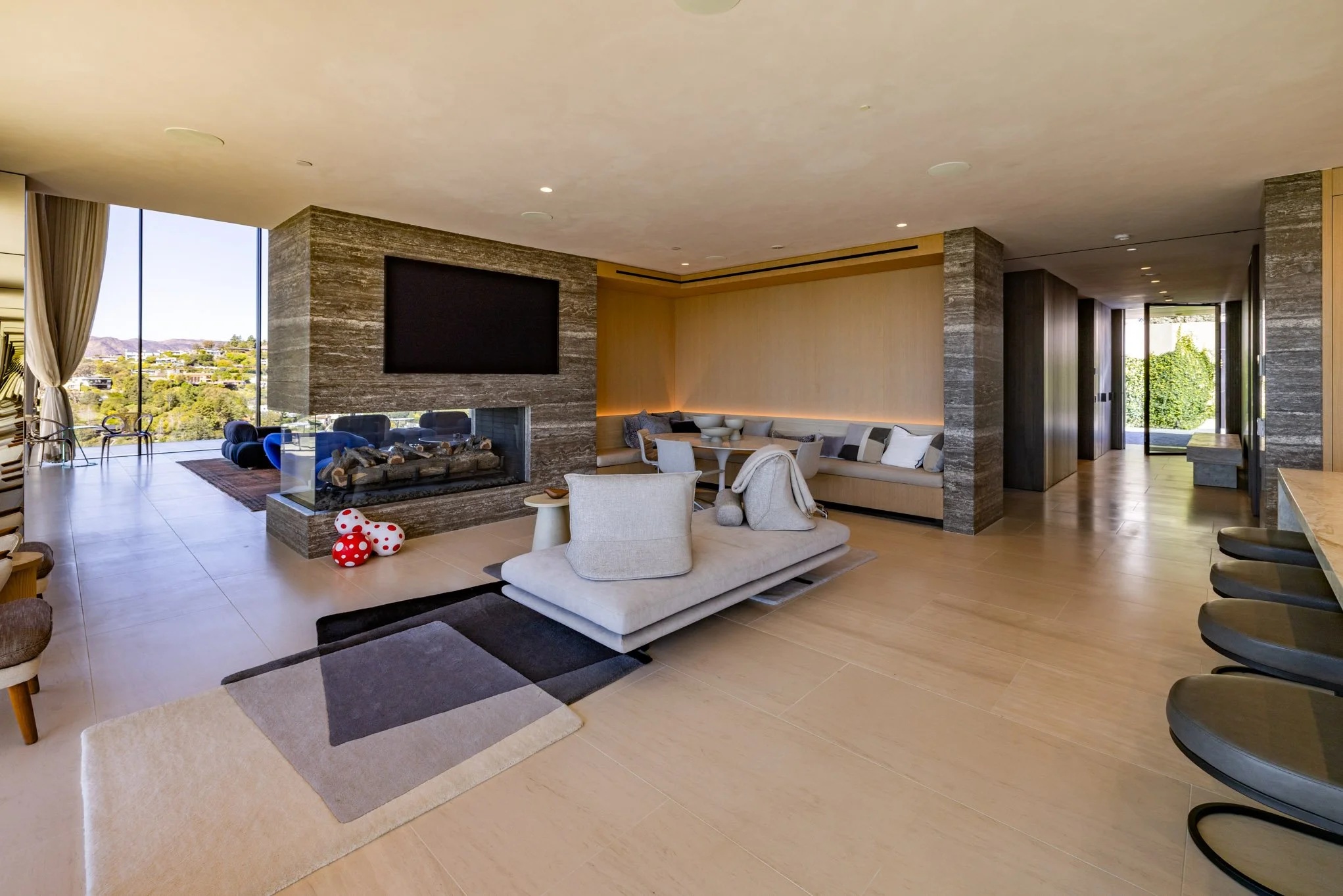 Modern living room with large windows, beige flooring, a fireplace with a TV, and contemporary furniture including a white sofa, beige armchair, and a sectional with pillows. There are bar stools on the right and an open hallway leading outside.
