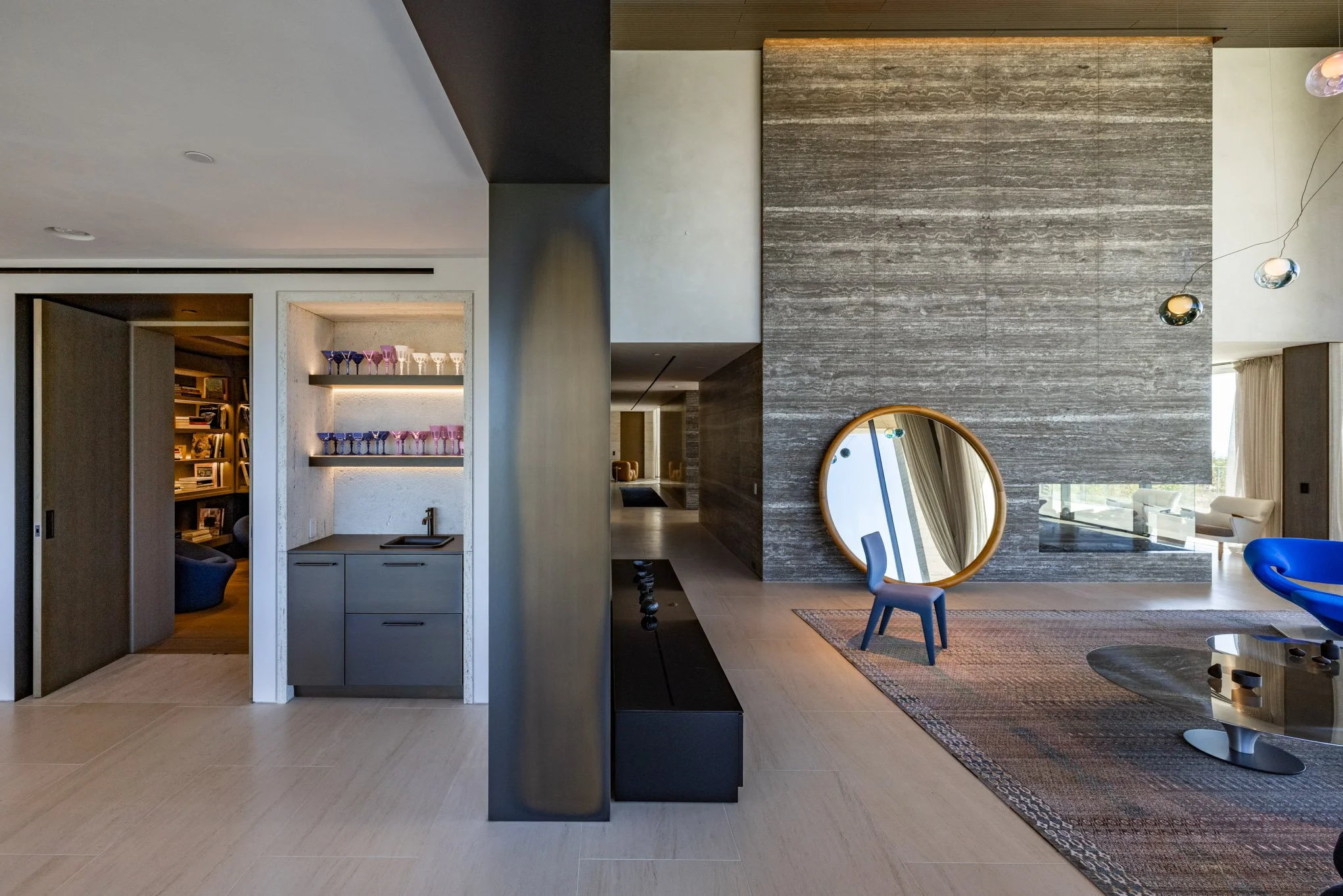 Modern living room with gray textured walls, circular mirror, colorful chairs, area rug, and built-in shelves with glassware.