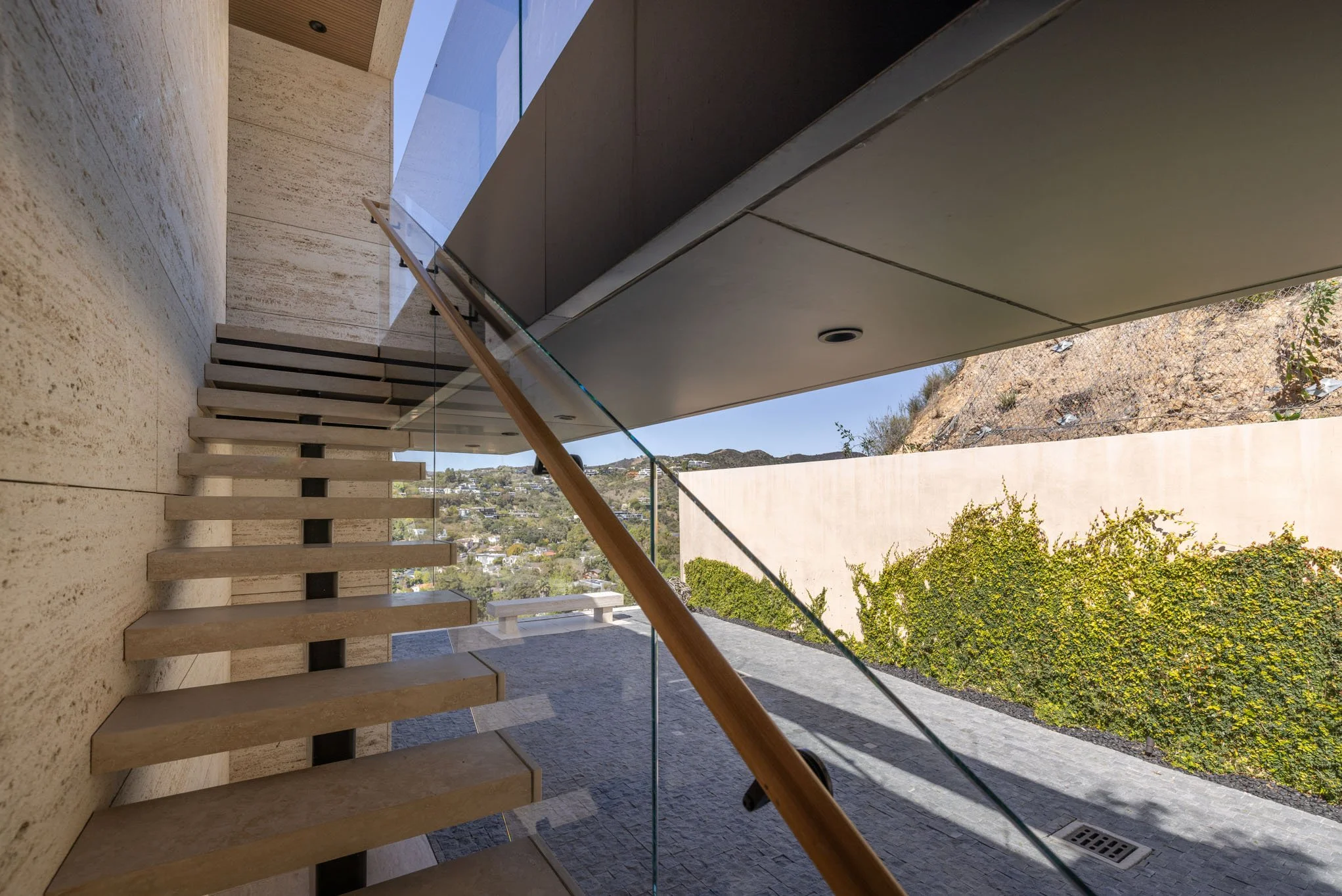 Modern outdoor patio area with stone steps, glass railing, and a landscaped background with green bushes and a hillside.