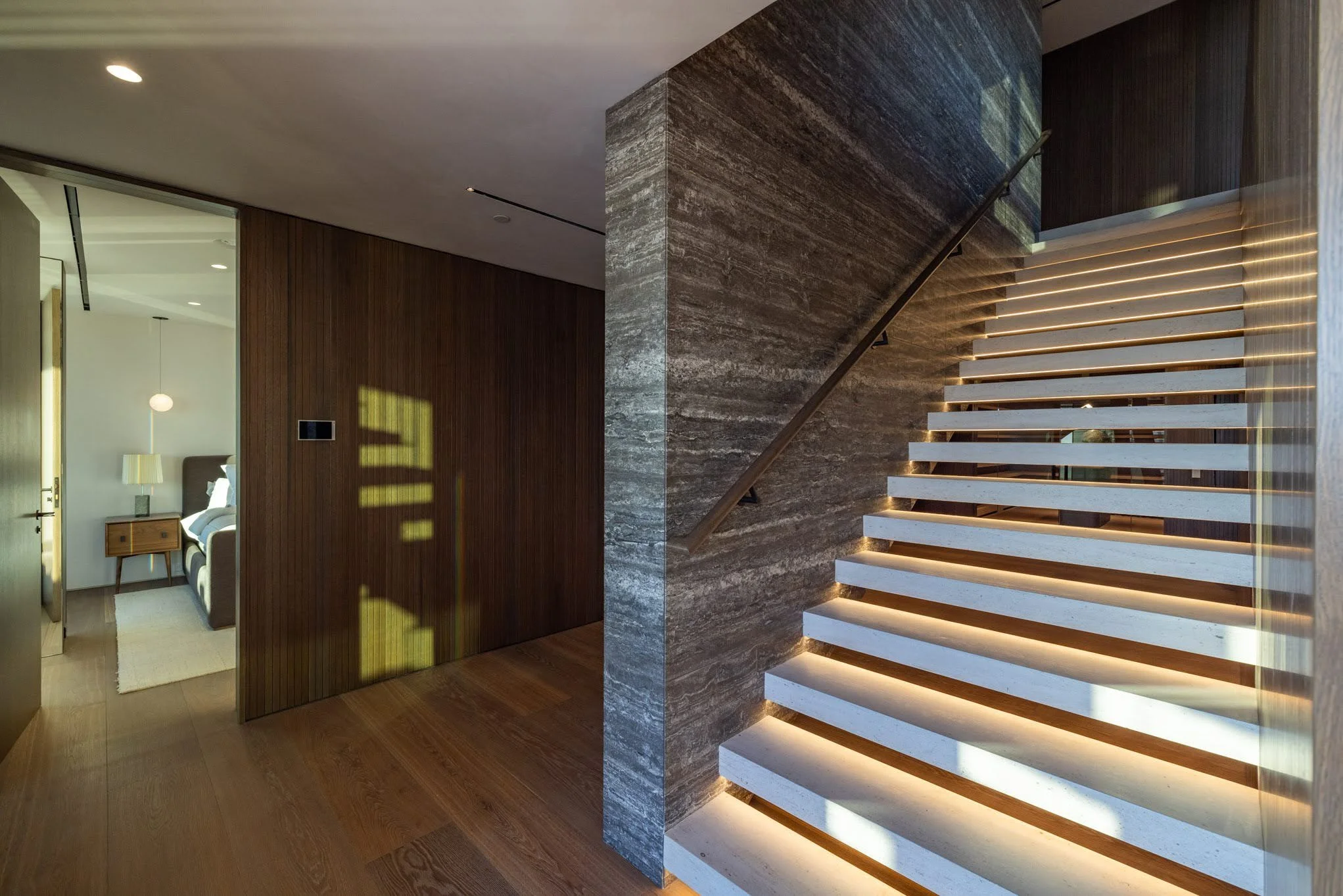 Interior view of a modern home with a staircase featuring LED lighting, wooden and marble walls, leading to upper levels, with a glimpse into a bedroom with a nightstand and lamp.