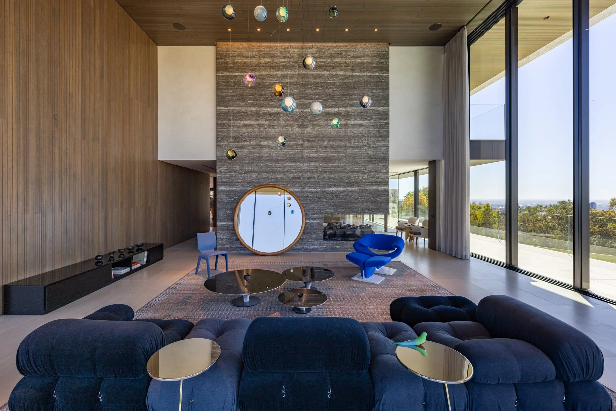 Modern living room with large floor-to-ceiling windows, dark velvet sectional sofa, blue accent chair, hanging pendant lights, circular mirrors, black and gold coffee tables, and a wooden-paneled wall.
