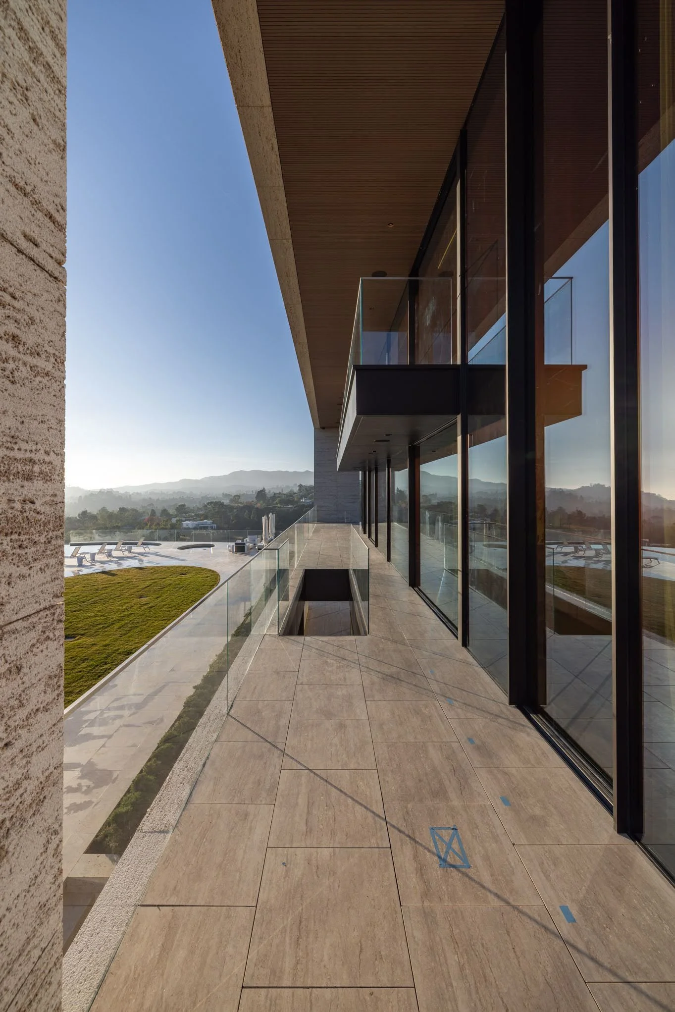 Modern balcony with glass railing, tiled floor, and large glass sliding doors, overlooking a landscape with hills and a swimming pool area.